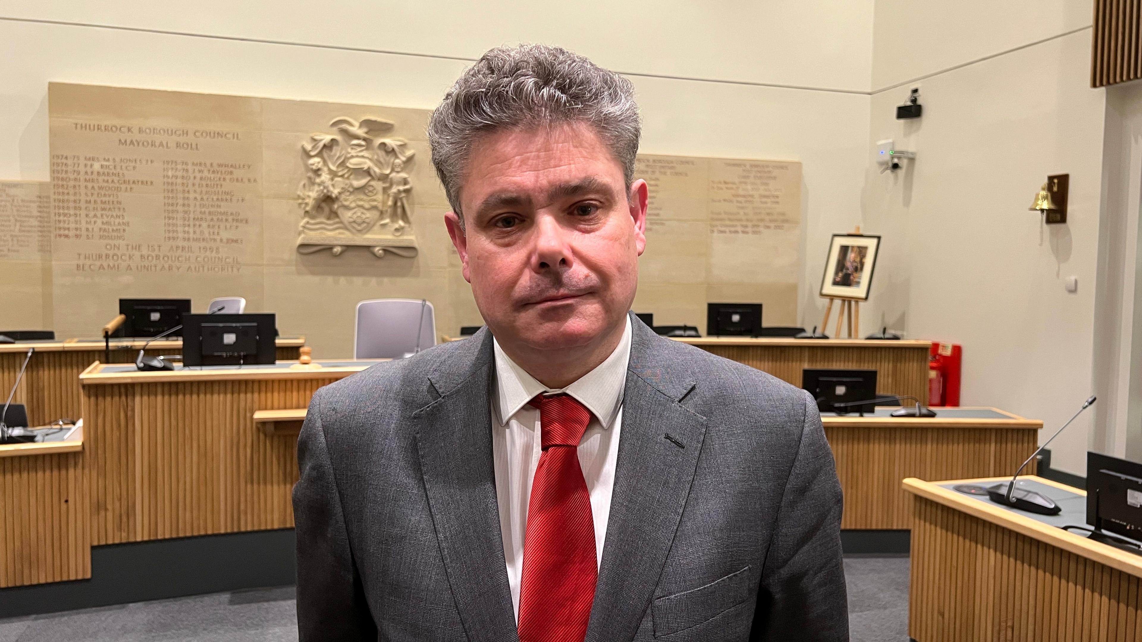 John Kent wearing a white shirt, red tie and grey suit jacket. He has grey hair. He is standing in the council chamber.