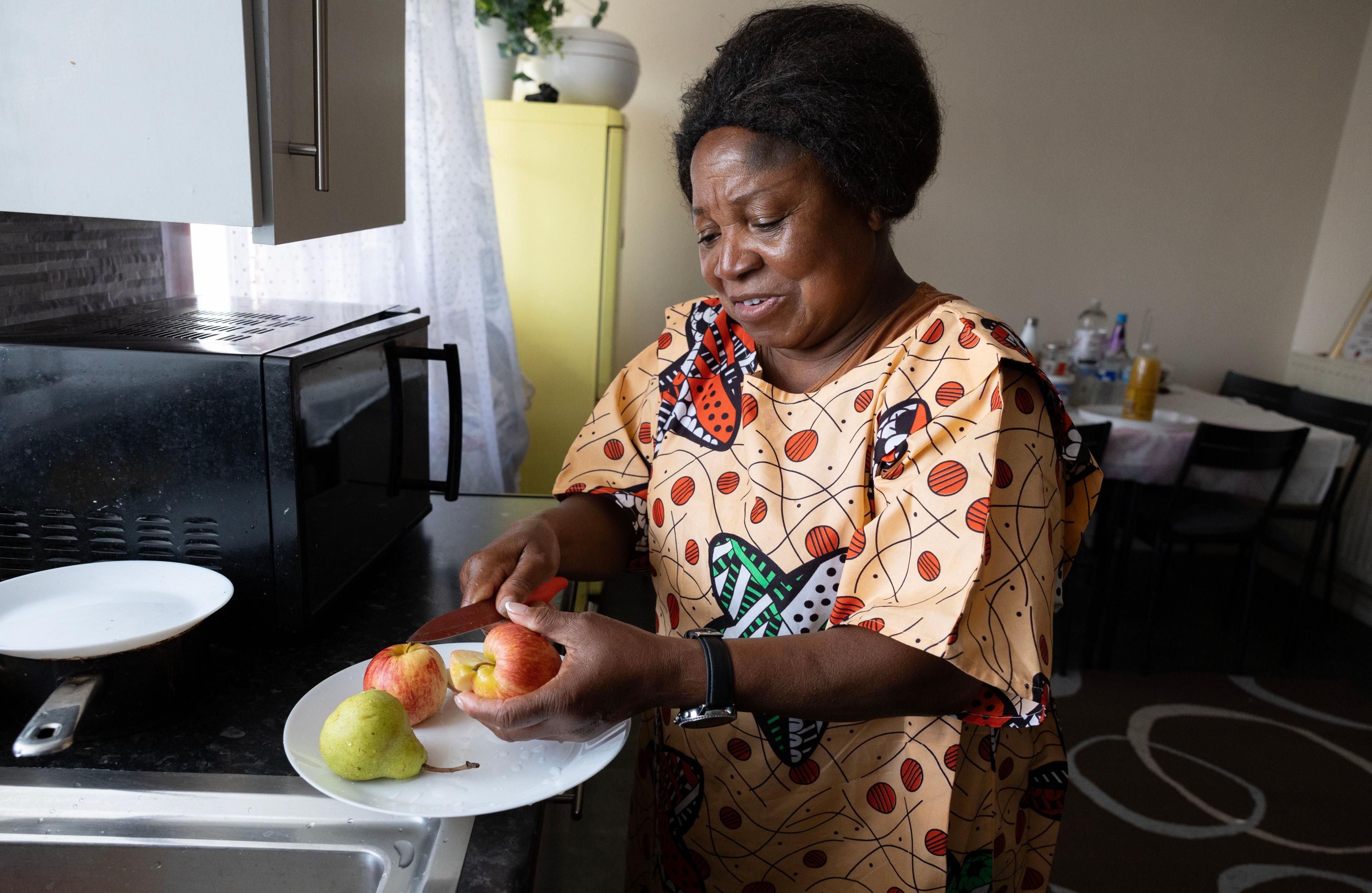 Rose chops two apples and a pear with a knife on a white plate in her home. She is wearing a colourful garment with butterflies