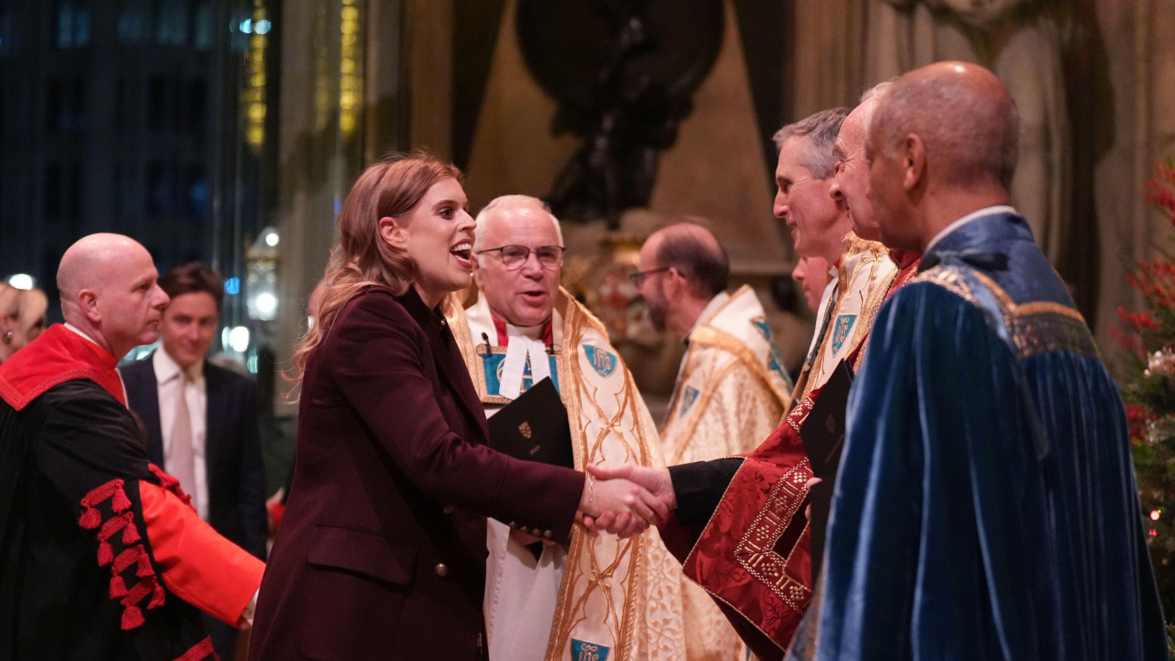 Princess Beatrice smiles as she shakes the hands of priests near the entrance. She is wearing a dark purple coat.