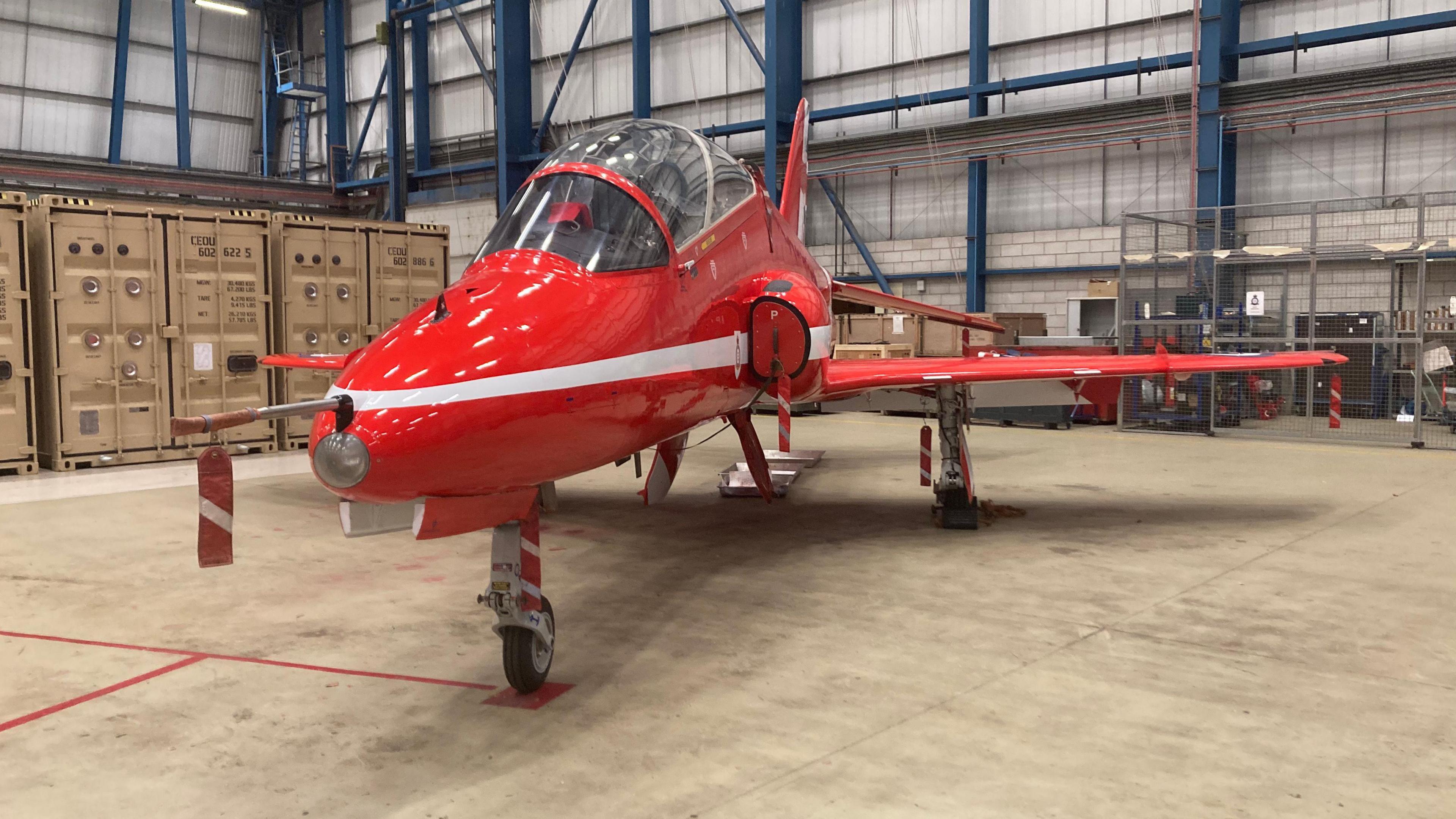 A stationary red aircraft situated in a warehouse building. The Hawk T1 is a fully aerobatic, low-wing, two-seat training aircraft.