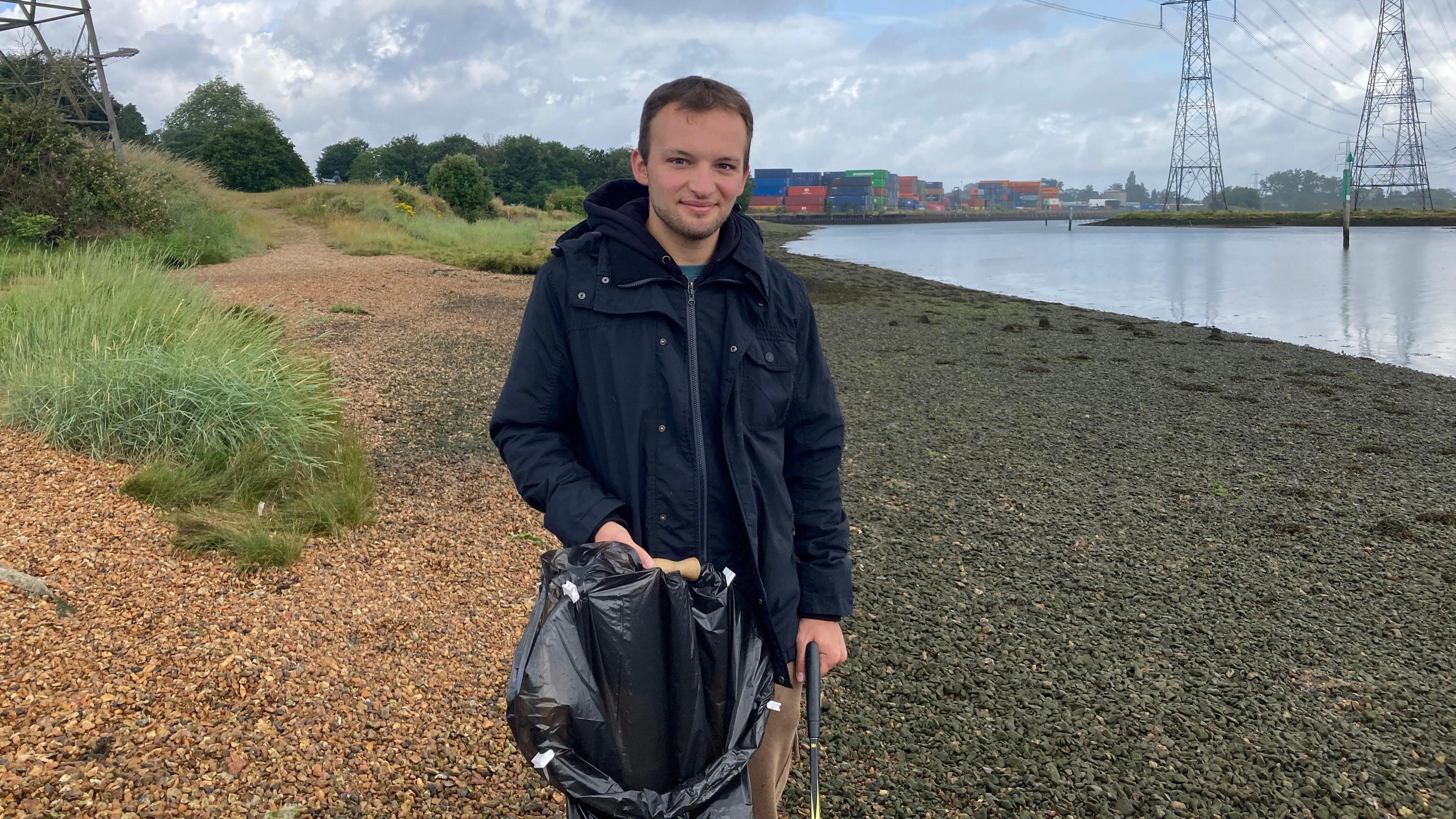 Dominik Reynolds stood on a beach holding a bin back in his hand and a stick used for picking up the litter