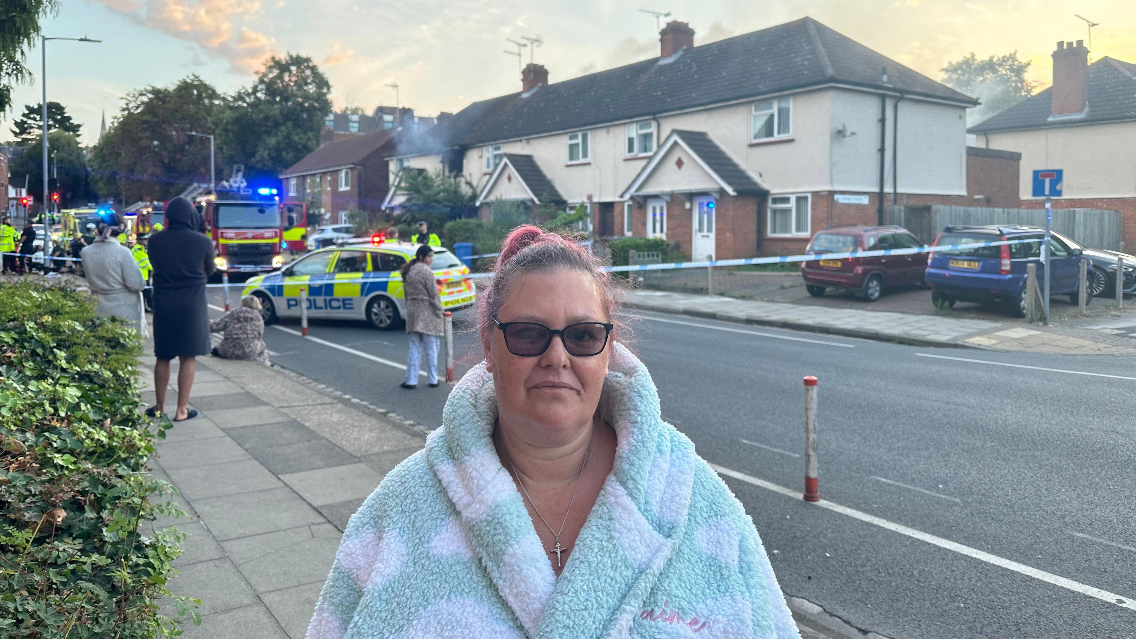 Resident Gina Gumble in green and white fluffy dressing gown and 
 black sunglasses, with the police cordon in the background. She is wearing a religious crucifix and has dark hair, in a ponytail with pick ends. 