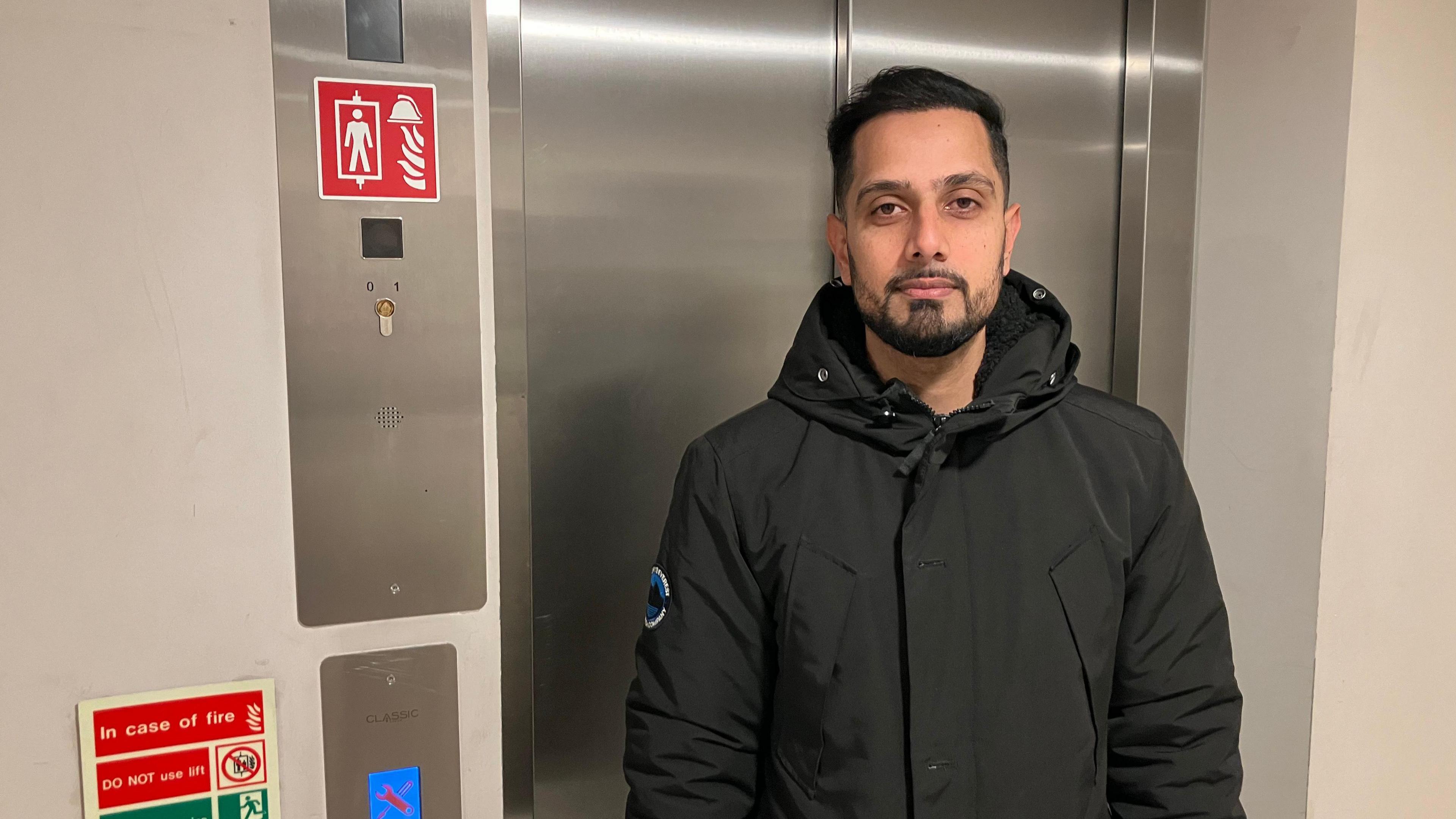 A man with dark hair and a beard wearing a black coat, standing in front of a set of lift doors and looking straight at the camera. On the wall next to him is a little screen with a picture of a spanner on it. 