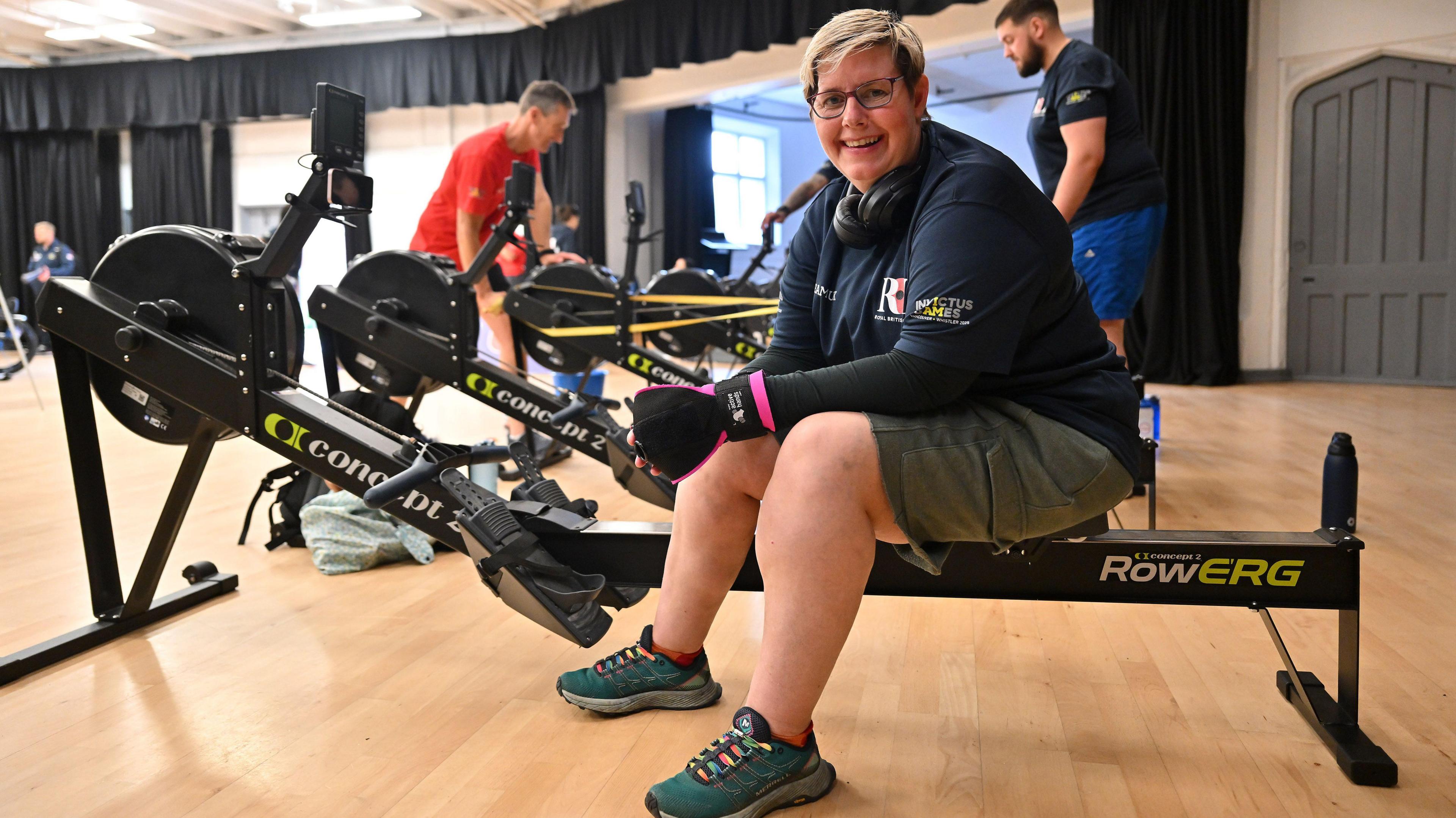 A woman with short blonde hair wearing gym gear has headphones around her neck. She is sitting on a rowing machine at the gym.