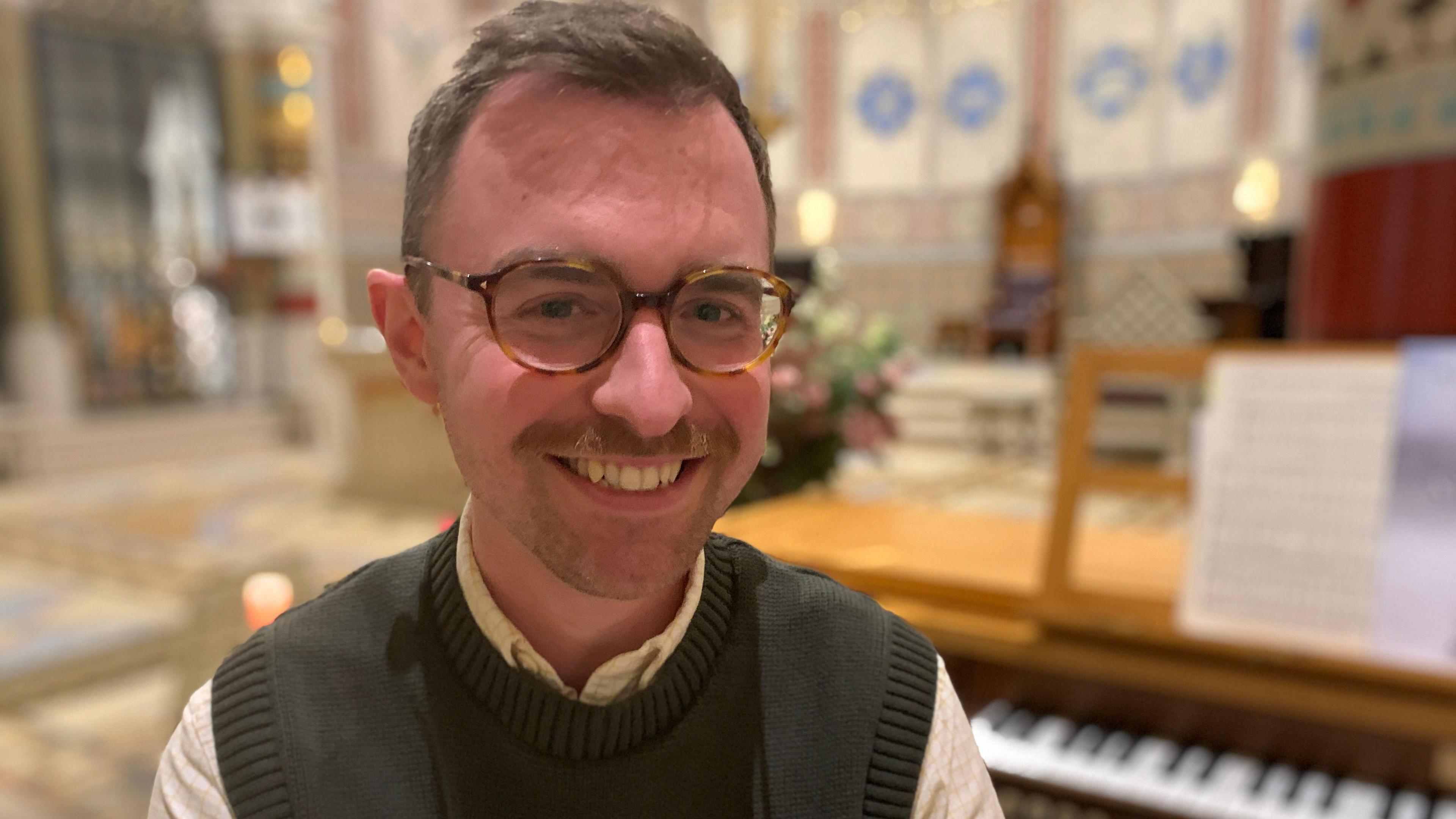 Image of James McConnell sitting in front of an organ. He has short brown hair and is wearing glasses, a cream shirt and a green knitted tank top.