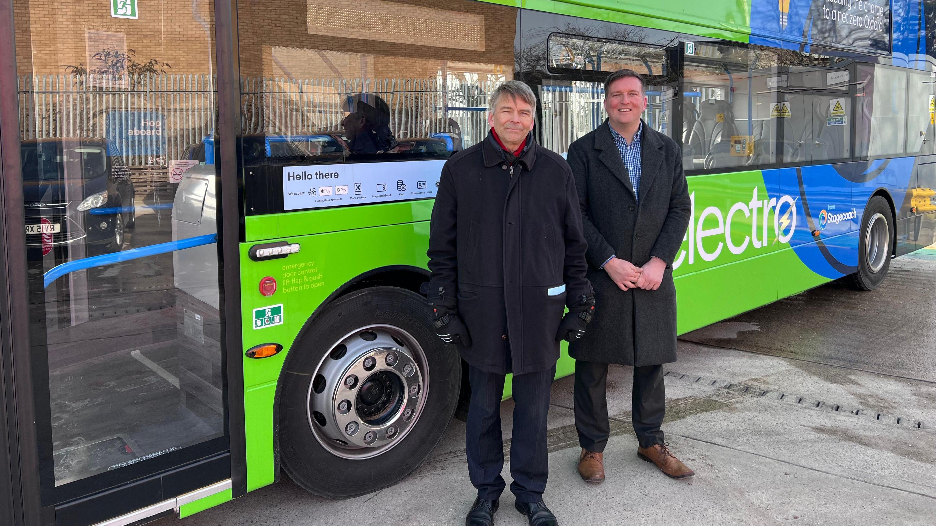 Councillor Andrew Gant and Stagecoach boss Chris Hanson stand together in front of a bus. Councillor Gant is on the left, he has short grey hair and wears a black collared wool coat, with a red collared jumper underneath and black smart trousers and black suit shoes and black hand mittens. Mr Hanson has short brown hair and wears a long collared wool coat with a blue and shite striped shirt and black smart trousers underneath with tan leather smart shoes. They are both standing in front of a green and blue electro bus.