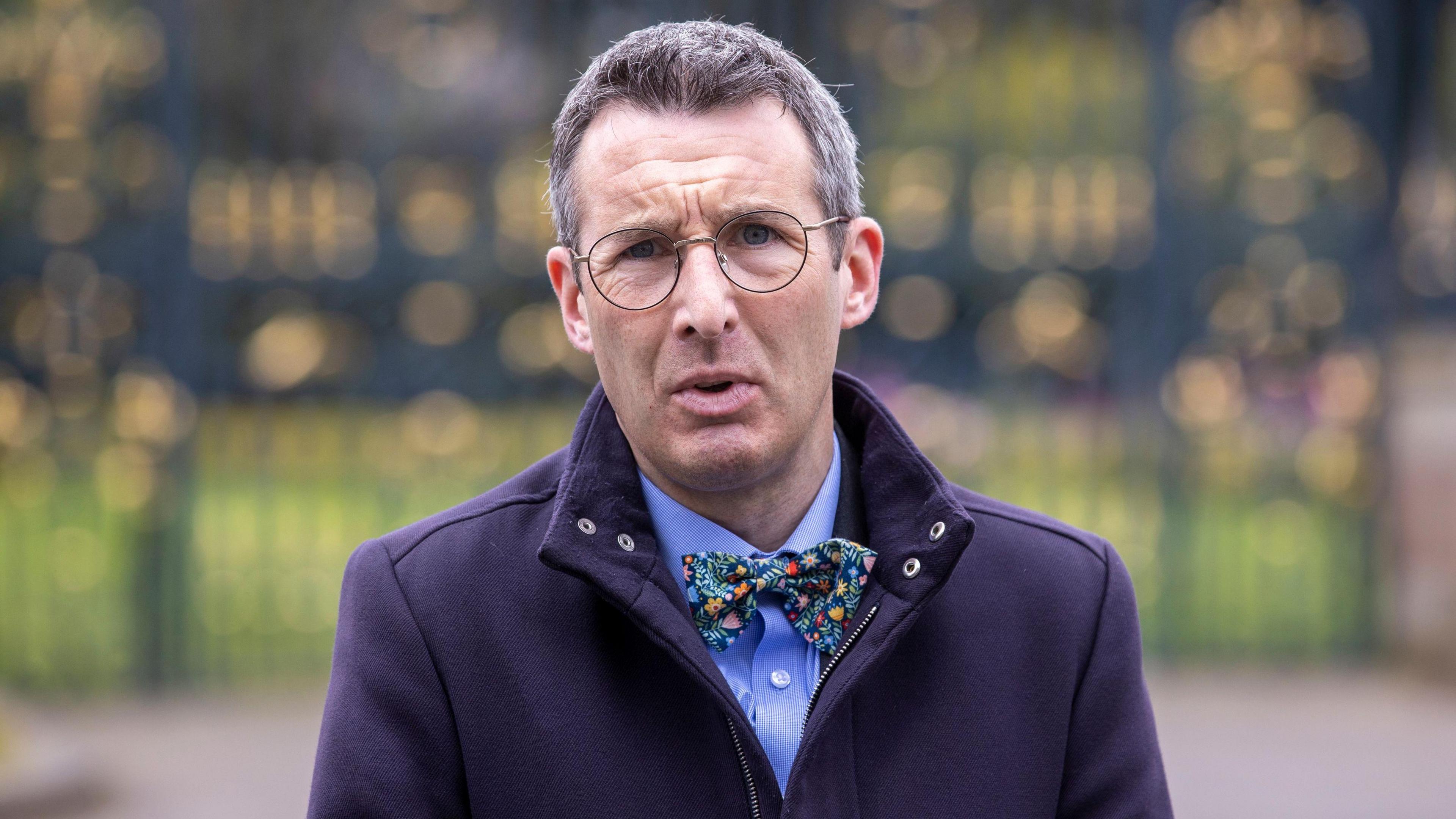 Andrew Muir wearing a coat, shit, and bowtie. He is speaking with a concerned expression on his face.