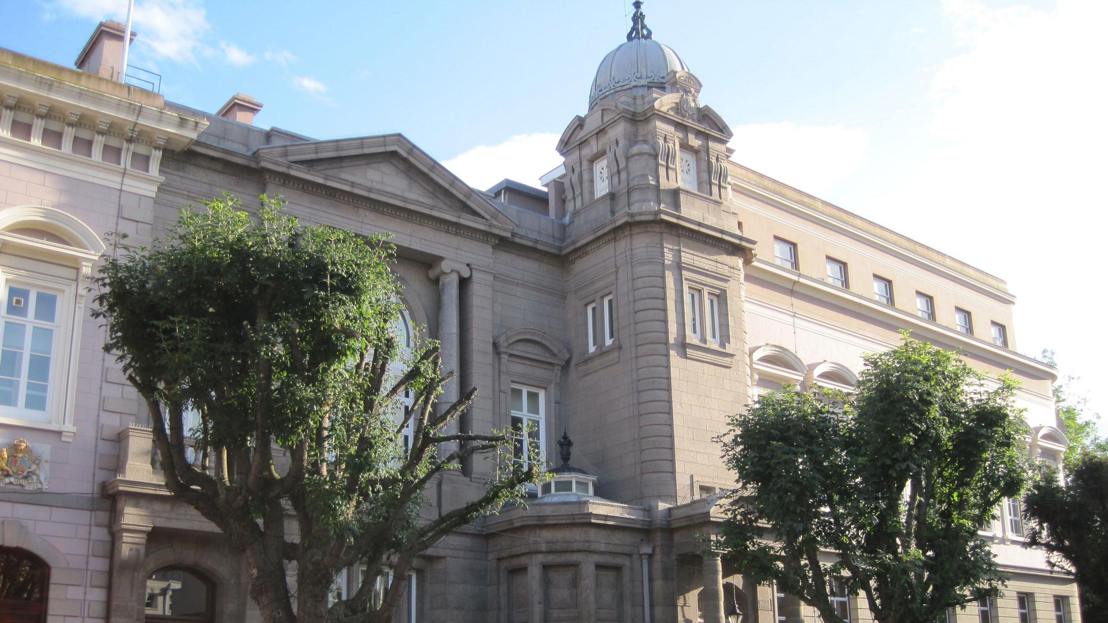 Royal Court in St Helier has a light grey exterior and has three storeys. The building appears to be from the Georgian era with long, thin windows. Three trees are positioned in front of the building.