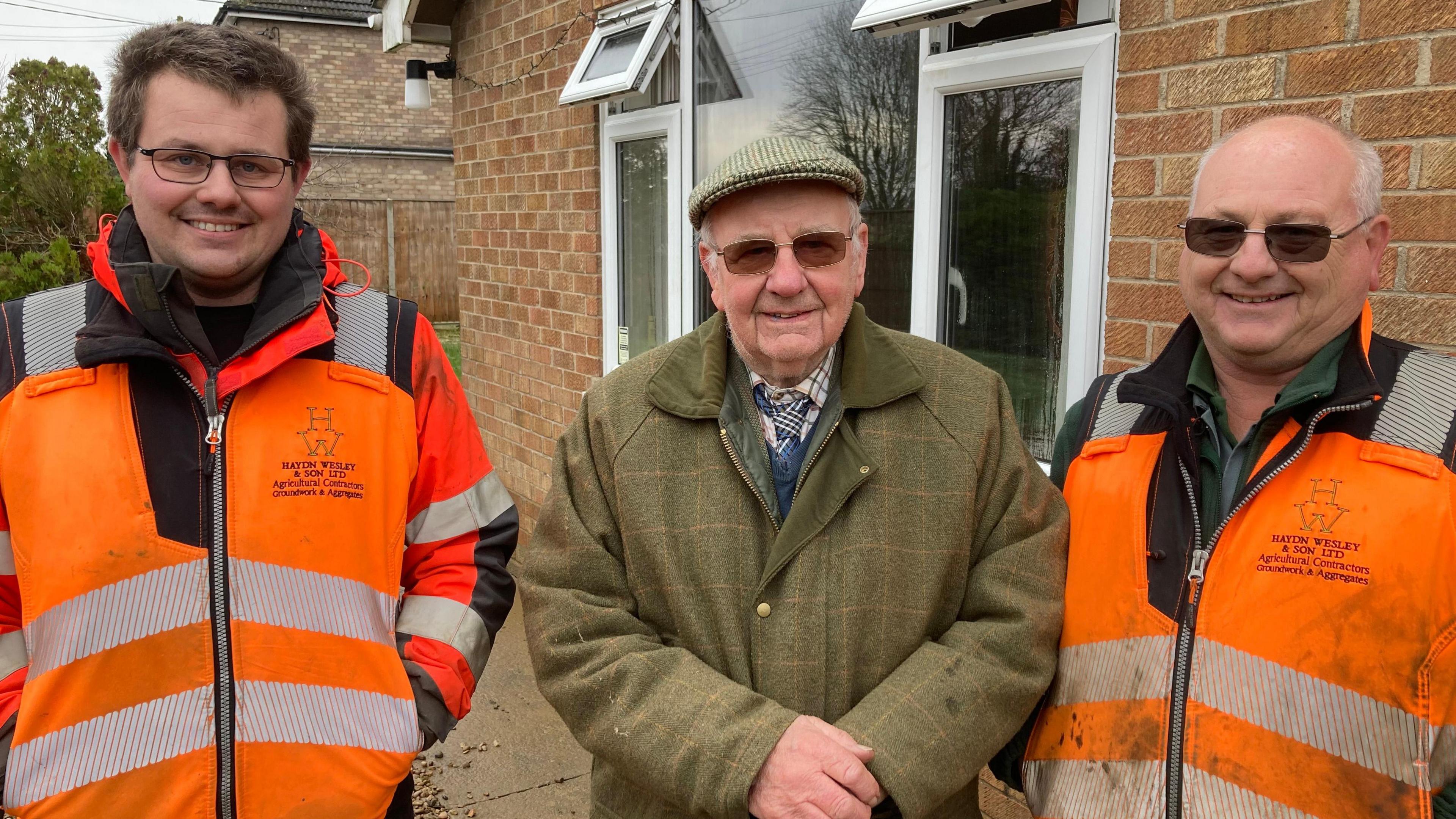 Three men stand in a row smiling and looking into the camera. Ryan Wesley (left) has short brown hair and is wearing black glasses and an orange hi-vis coat. Hayden Wesley (middle) is wearing a tweed cap, glasses and a tweed coat. Dean Wesley (right) has short grey hair and is wearing an orange hi-vis coat. 