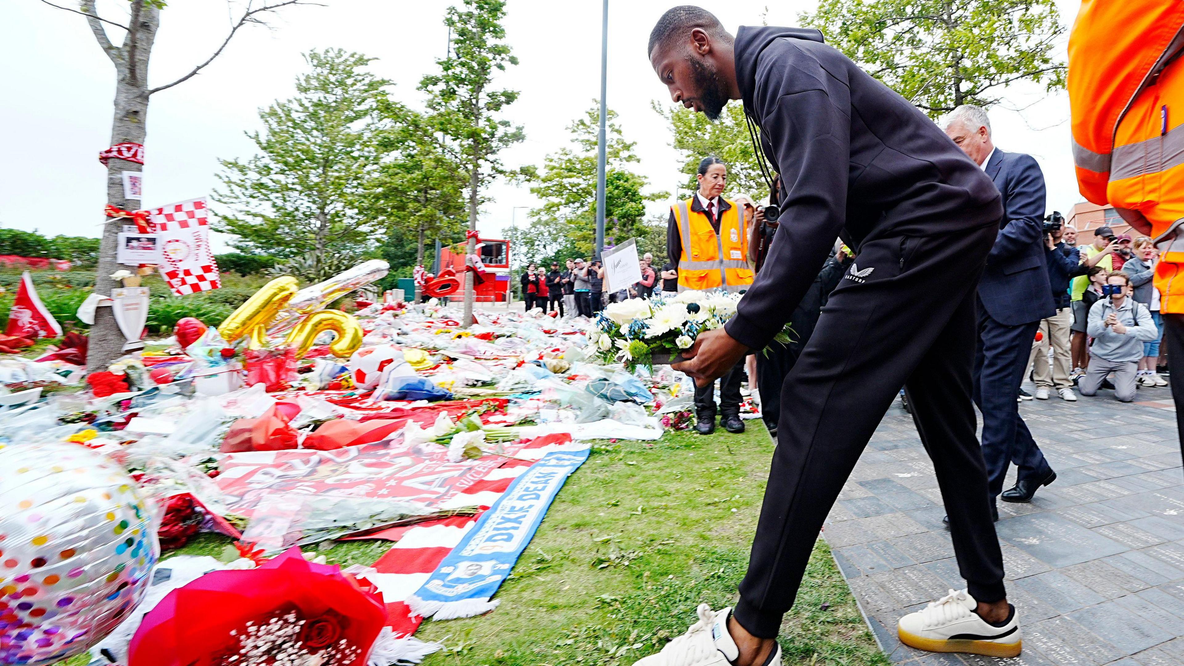 Beto, in a navy blue tracksuit, places a wreath made up of white flowers on to a grassy area containing colourful tributes including bouquets, scarves, balloons and flags.