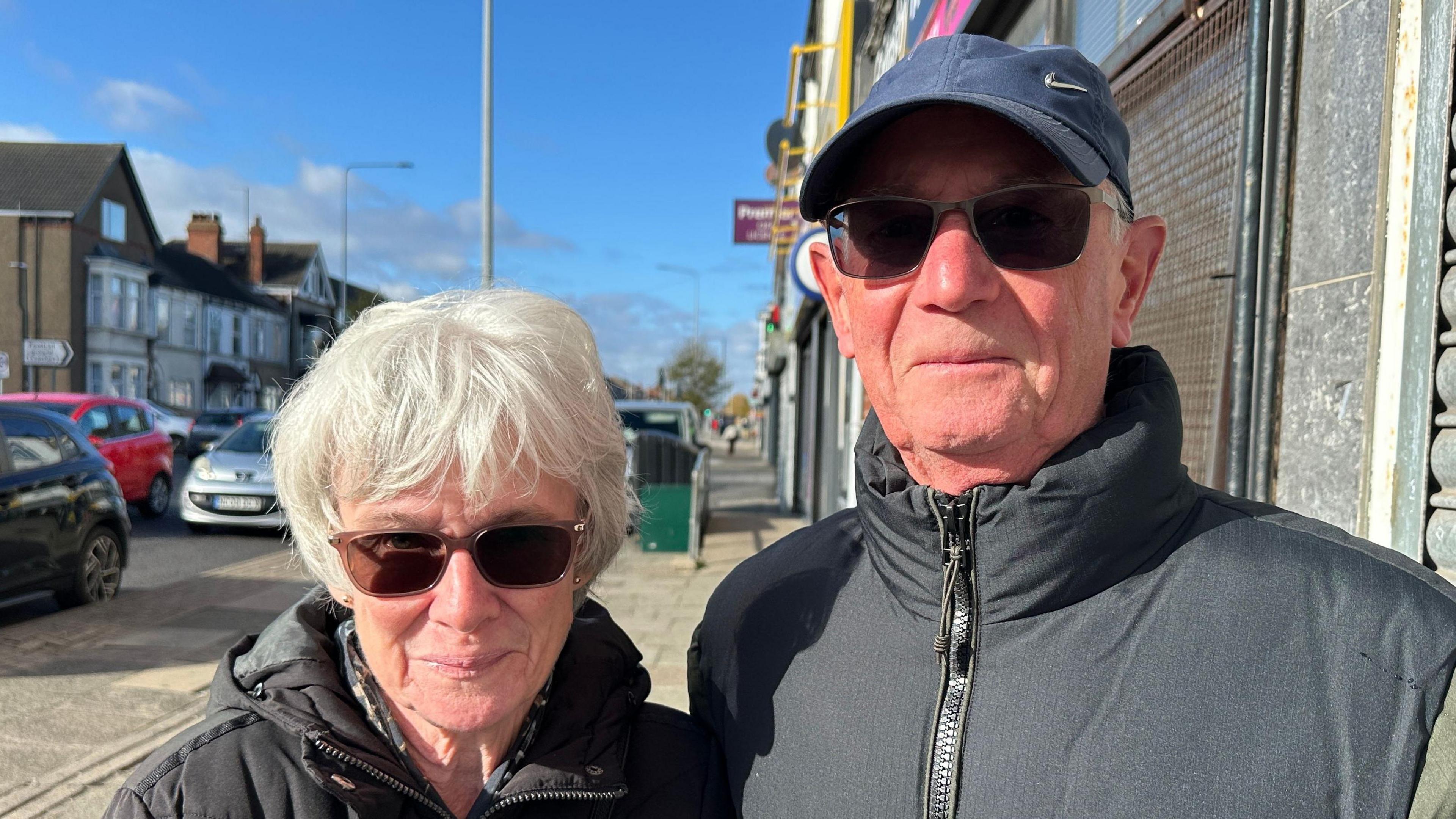 Val and Mick Carr stood in the street smiling at the camera. Val Carr has short grey hair and is wearing brown sunglasses and a black hooded coat. Mick Carr is wearing a navy Nike cap, black sunglasses and a black bodywarmer. The sky is blue. 