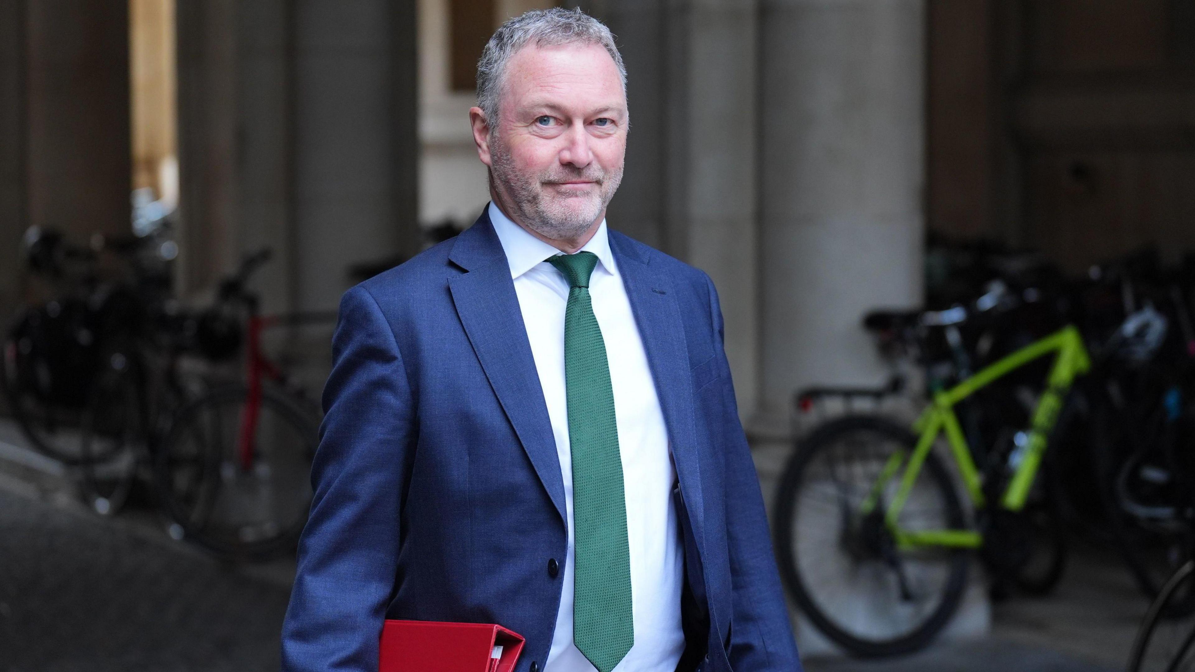 Steve Reed looks at the camera as wearing a navy suit with white shirt and green tie. He is carrying a red folder. He is walking through a passageway with bikes parked in the background.