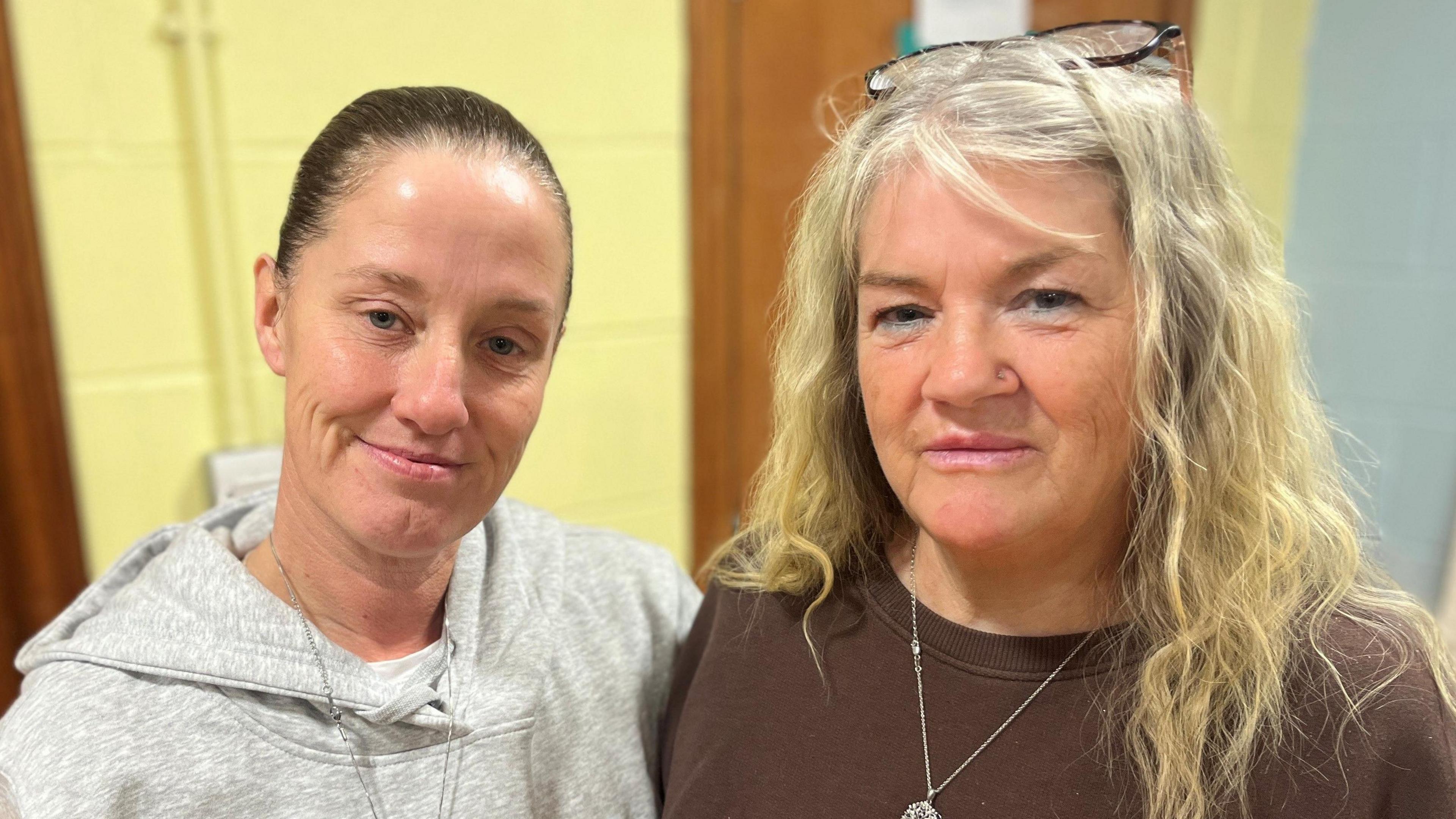 Lisa Petrie has her dark hair tied up and wears a grey hoodie. She has her arm around Pauline Bowie, a woman with long blonde hair wearing a brown jumper.