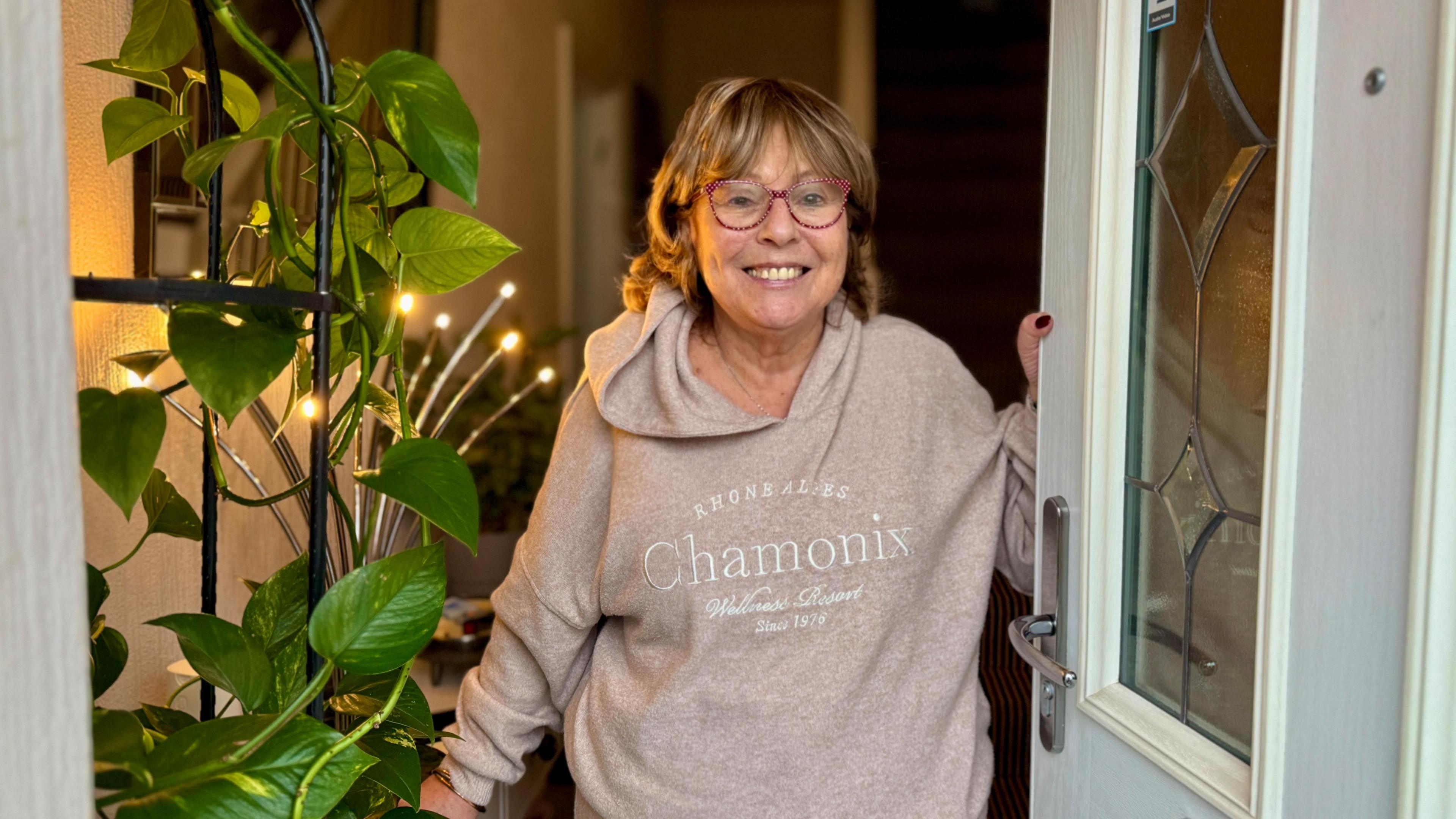 A woman wearing a brown hood is stood smiling at her front door. She has a green plant next t her and is lit from behind by a warm light.