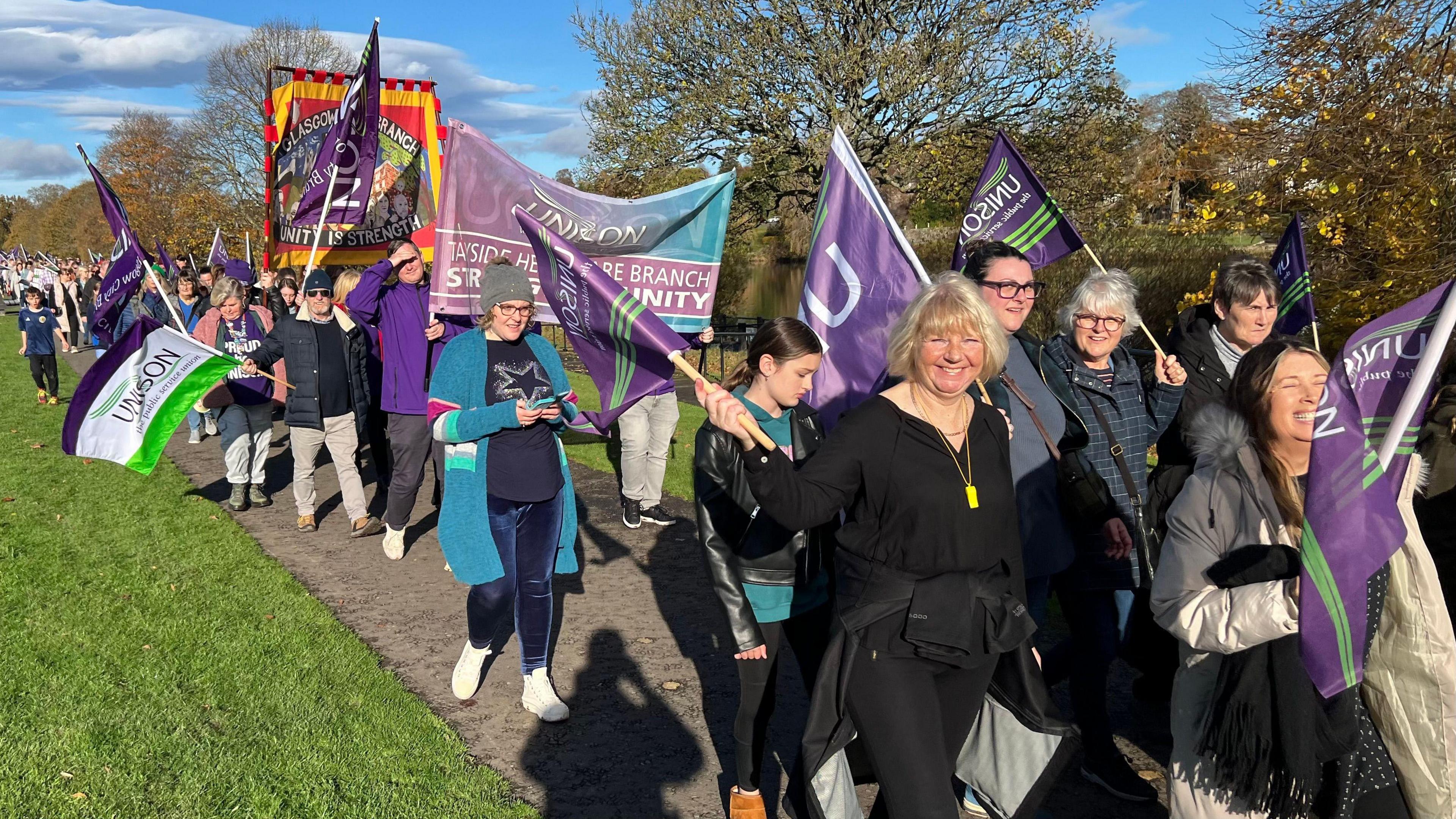 A group of people on a march in a park, waving purple Unison flags