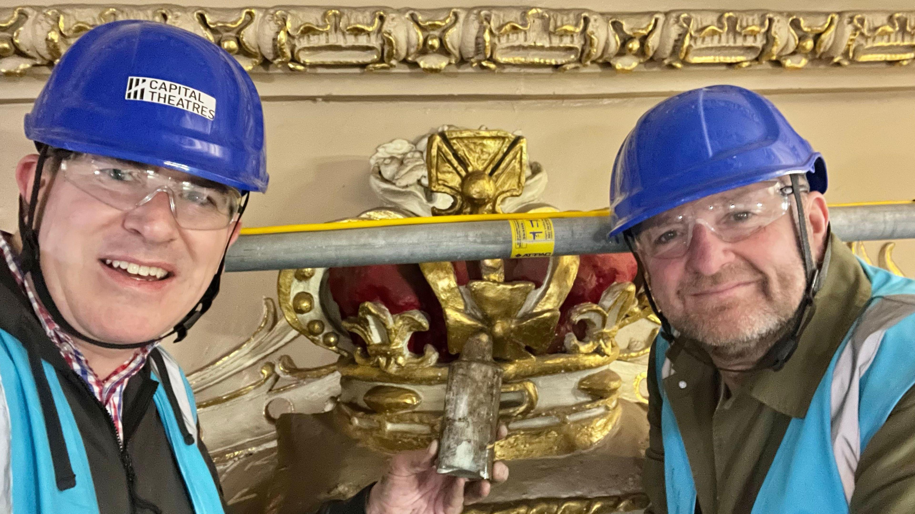 Mike Hume and John Robb wearing blue helmets and safety goggles. They are beside the crown which is painted gold and maroon. There is a scaffolding pole as they are on a raised platform.