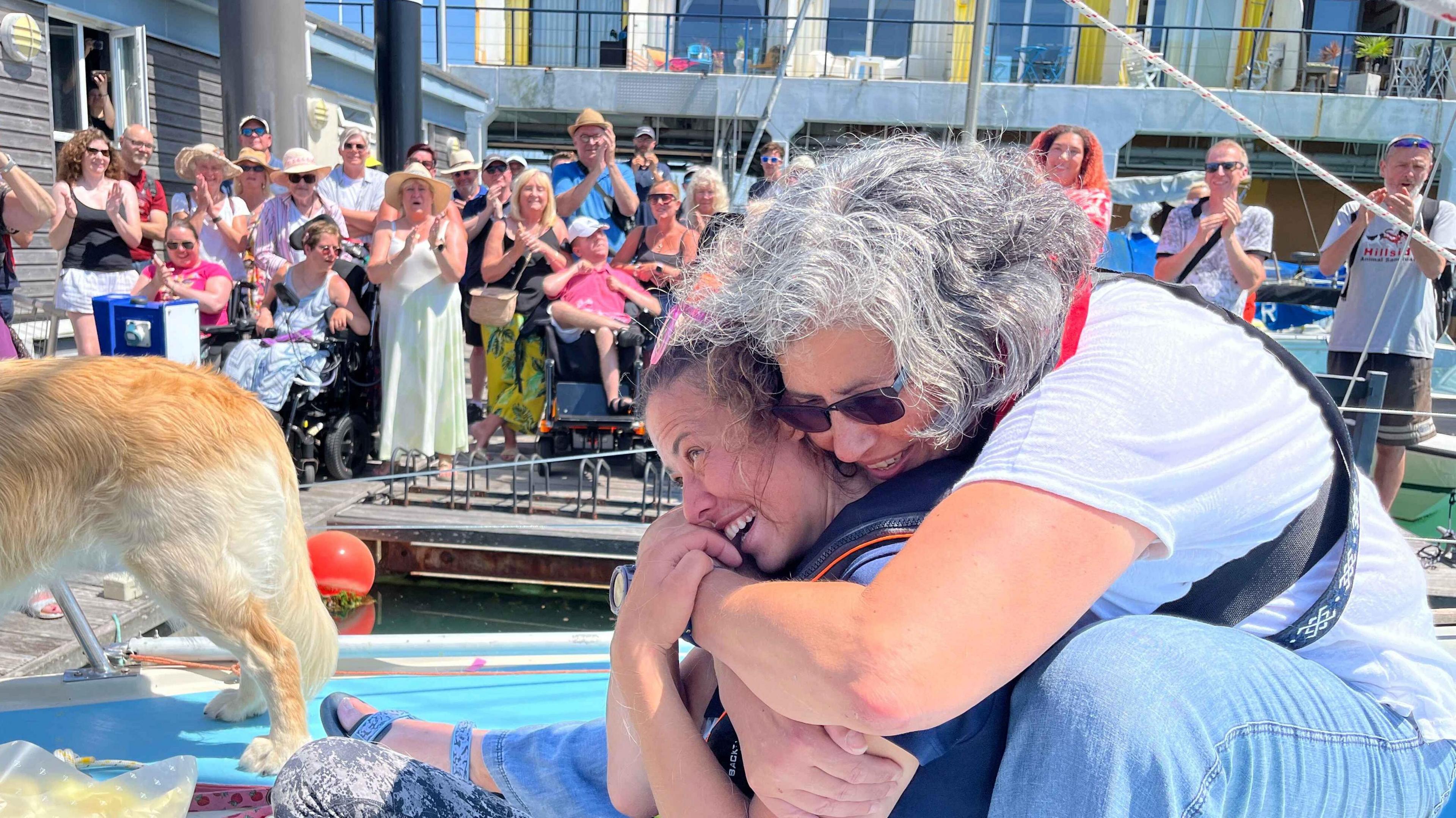 Jazz sat on the front of her boat being hugged by a family member with a huge crowd cheering her on the jetty in the background
