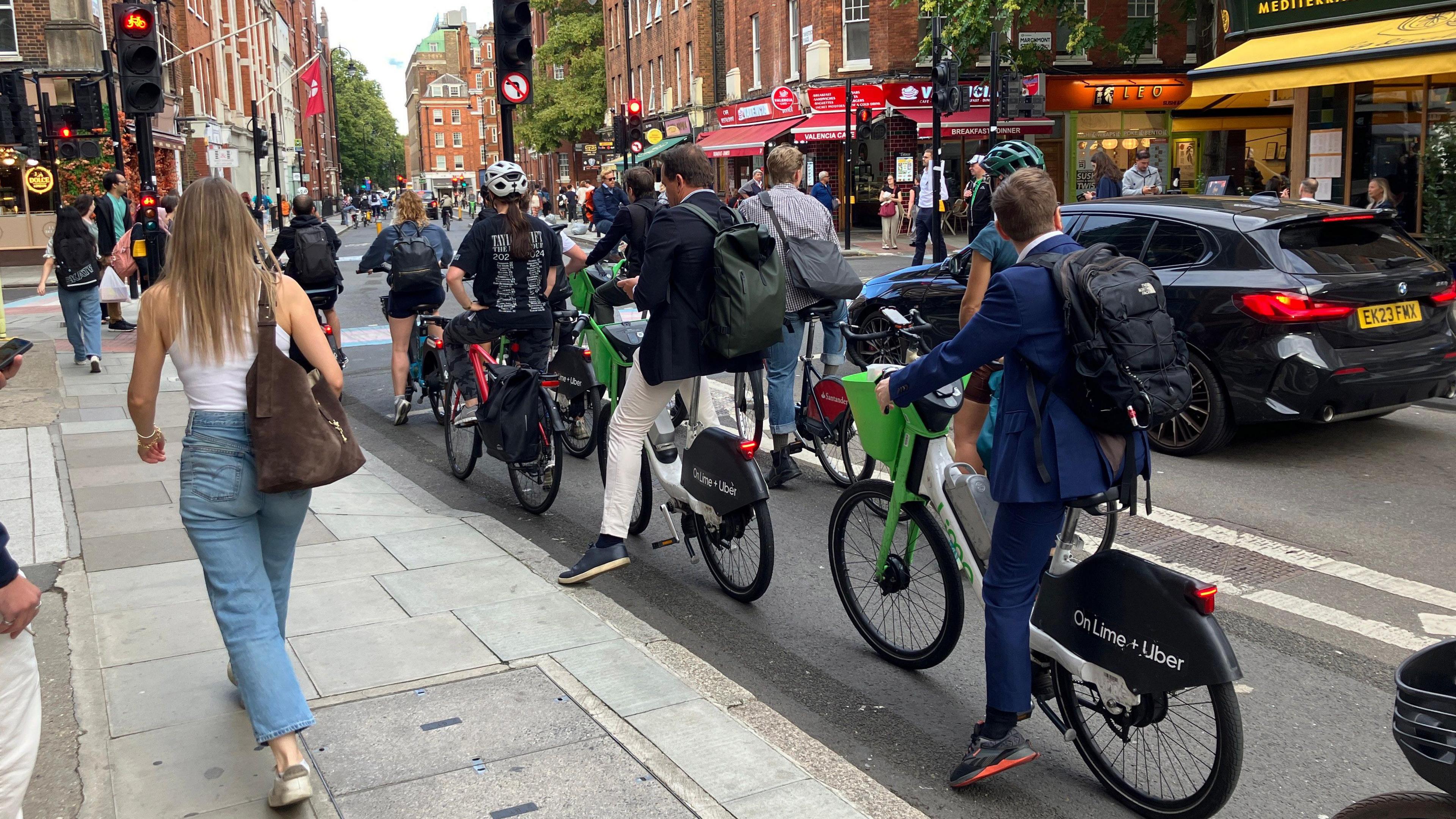 Cyclists, including Lime Bike users, wait at a red traffic light as pedestrians walk past