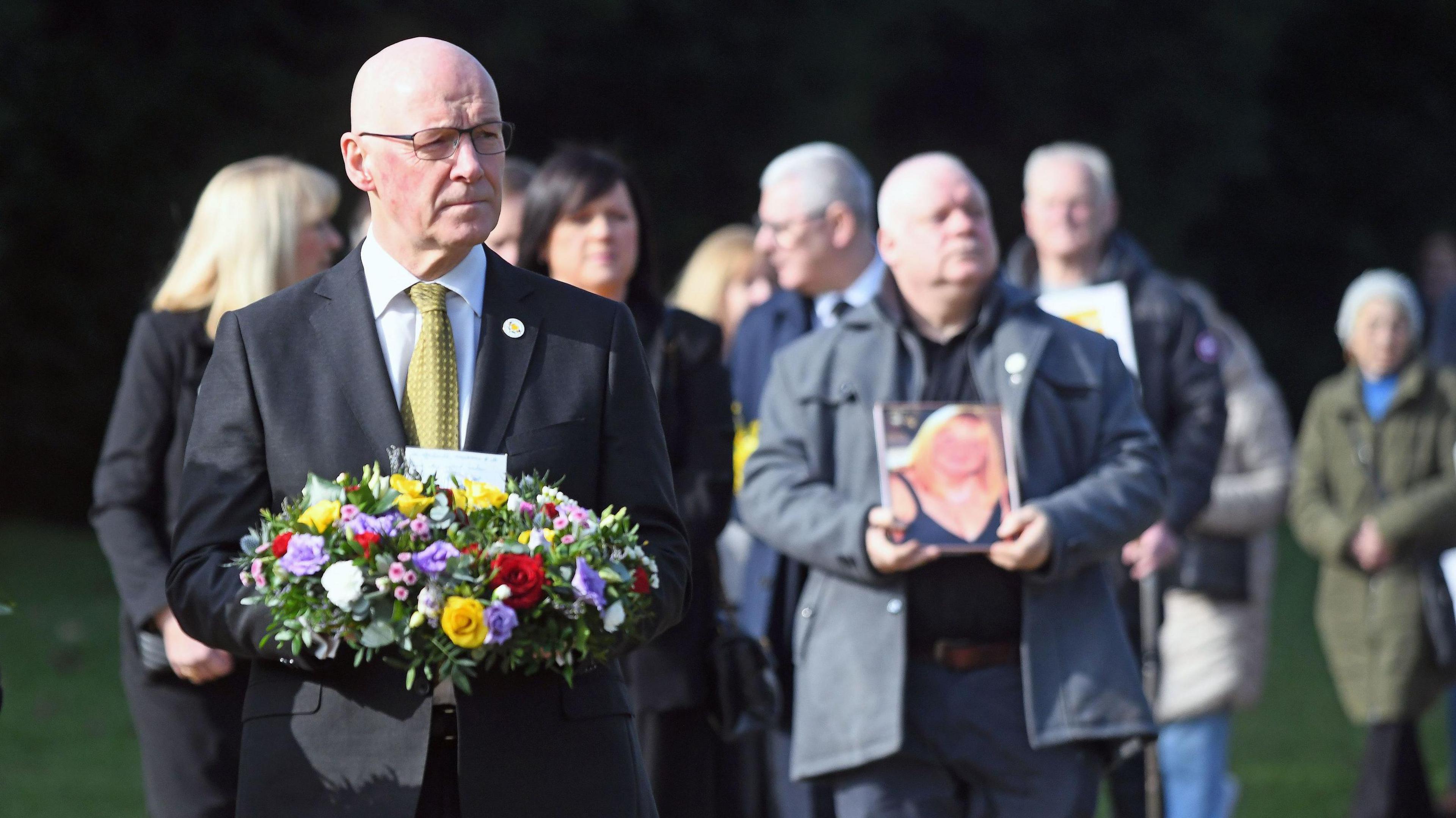 Swinney wearing a dark suit and yellow tie carries a multicoloured wreath of flowers as people walk behind him.