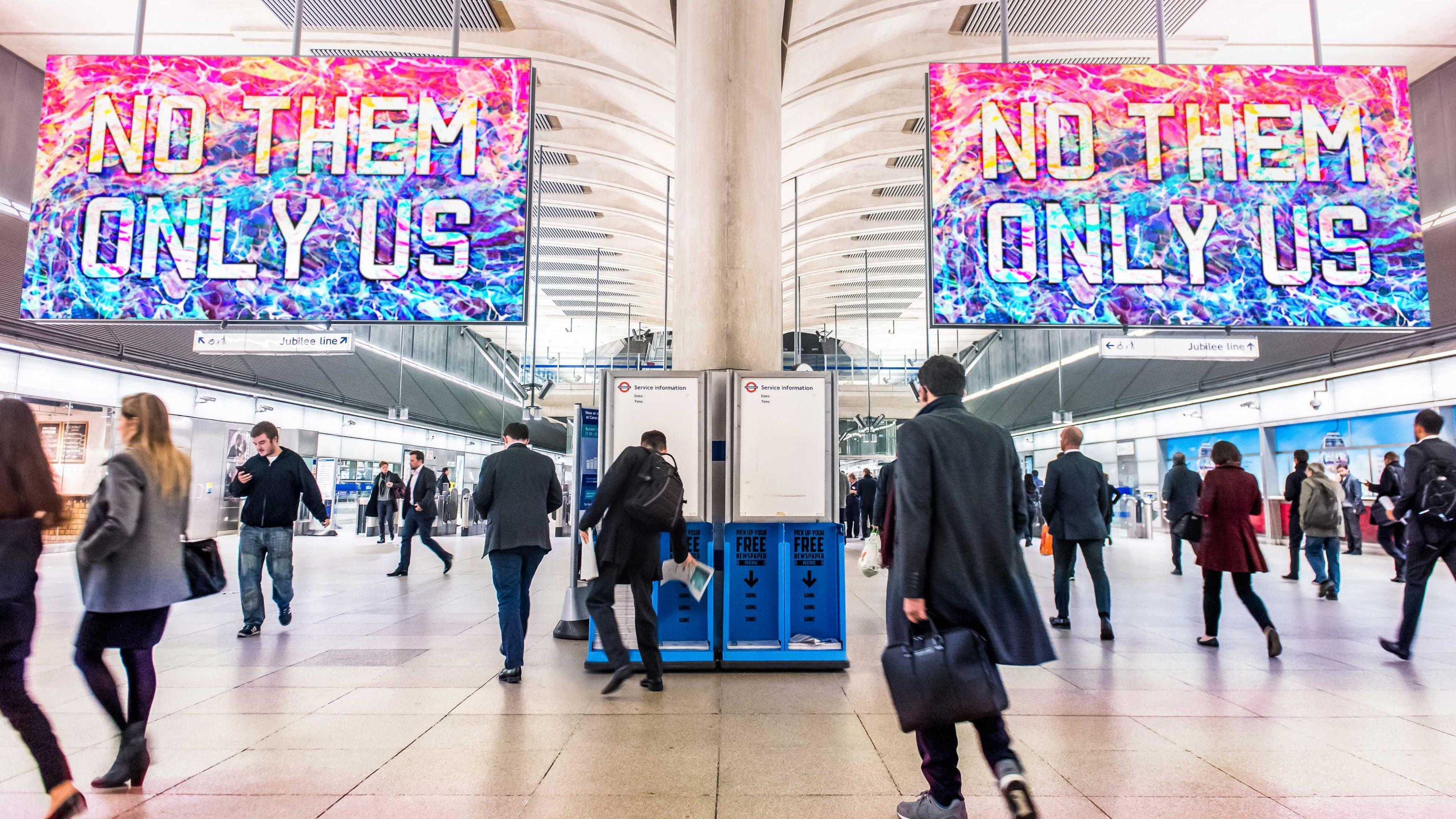 Two large boards sit above a busy Tube station concourse. They both read "No them only us" in white capital letters against a stained glass effect background of mostly blues and purples. 