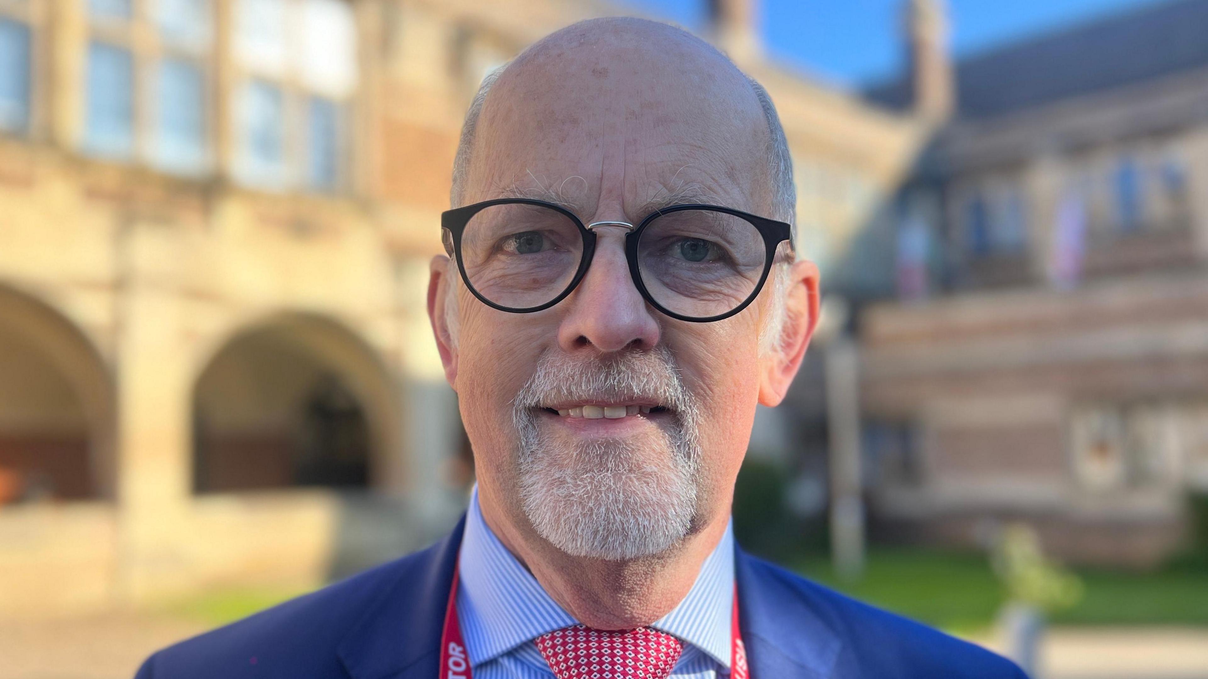 A man with glasses and a white beard smiles at the camera. He is standing in front of the school and wears a blue suit jacket, blue stripe shirt and red checked tie. He is wearing a red lanyard.