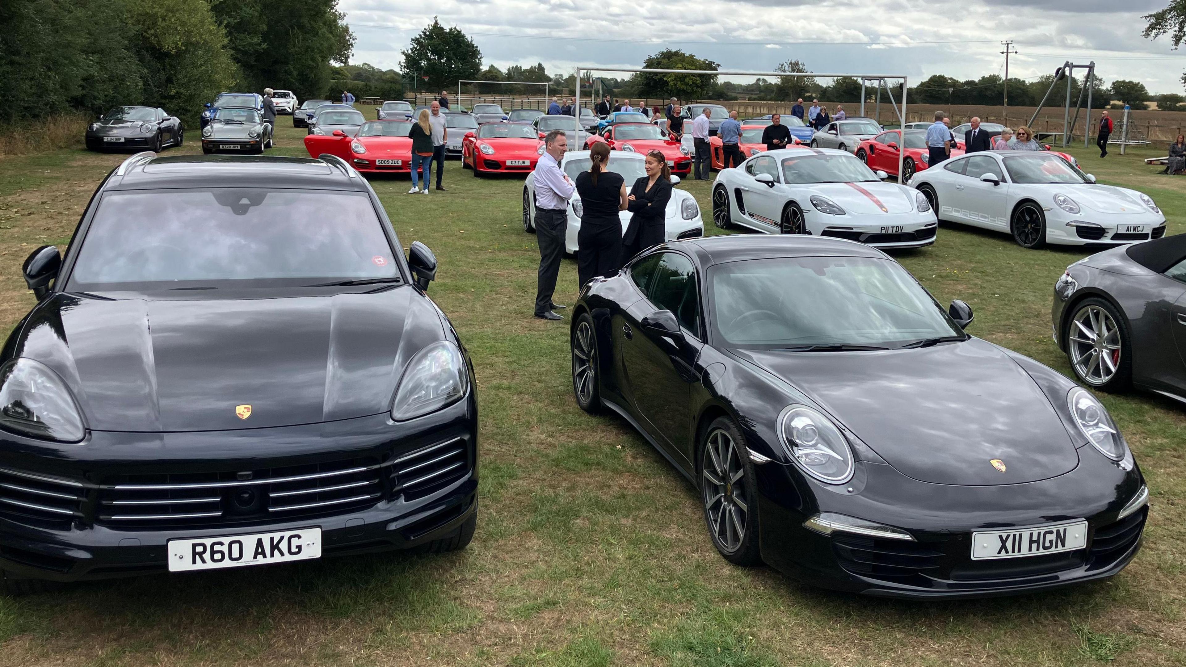 Two black Porsches are at the front of the image, with other colours of Porsches lined up behind. They are sat on a field.