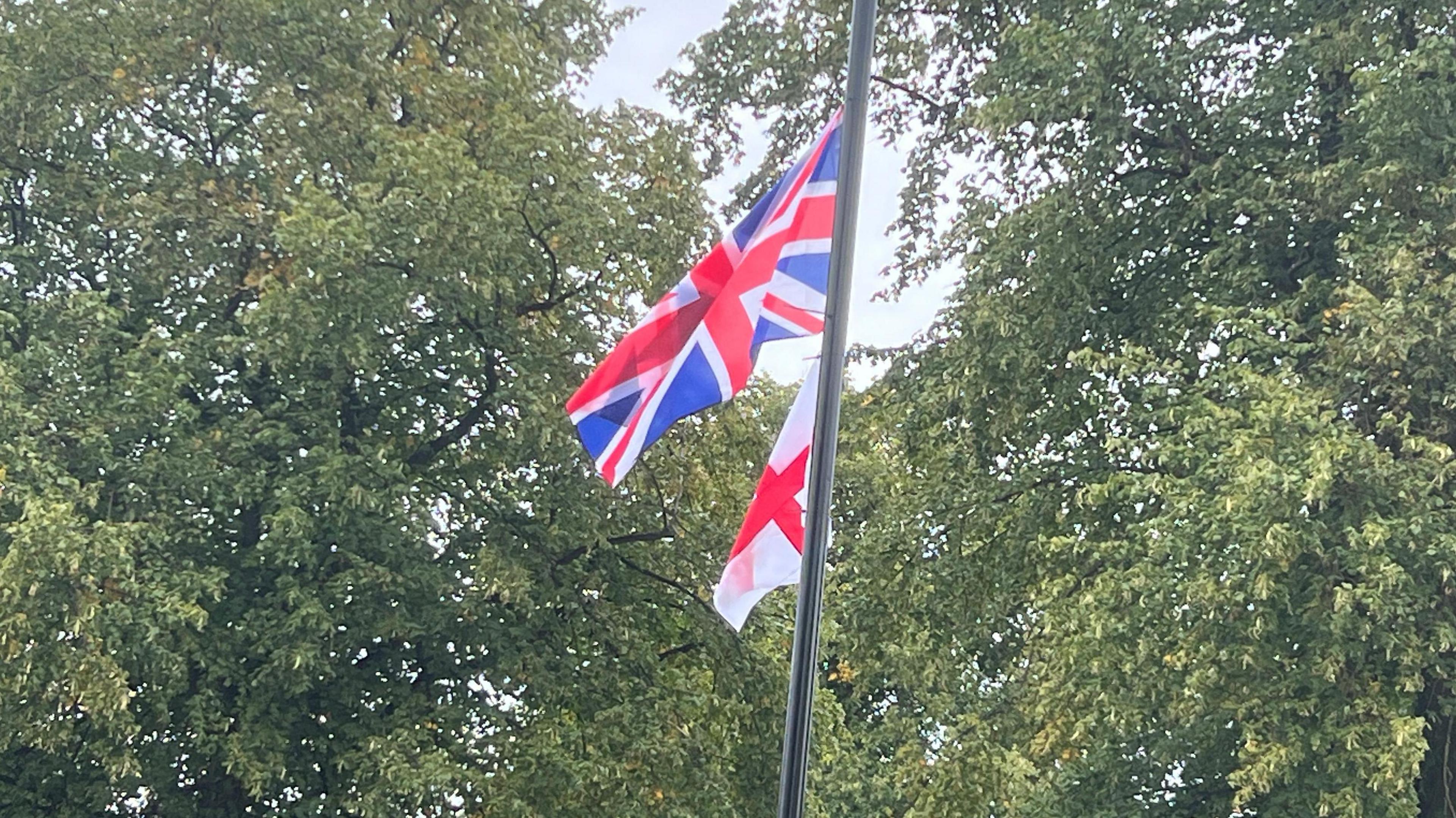 A lamppost with a Union flag and England flag attached, and trees in the background.