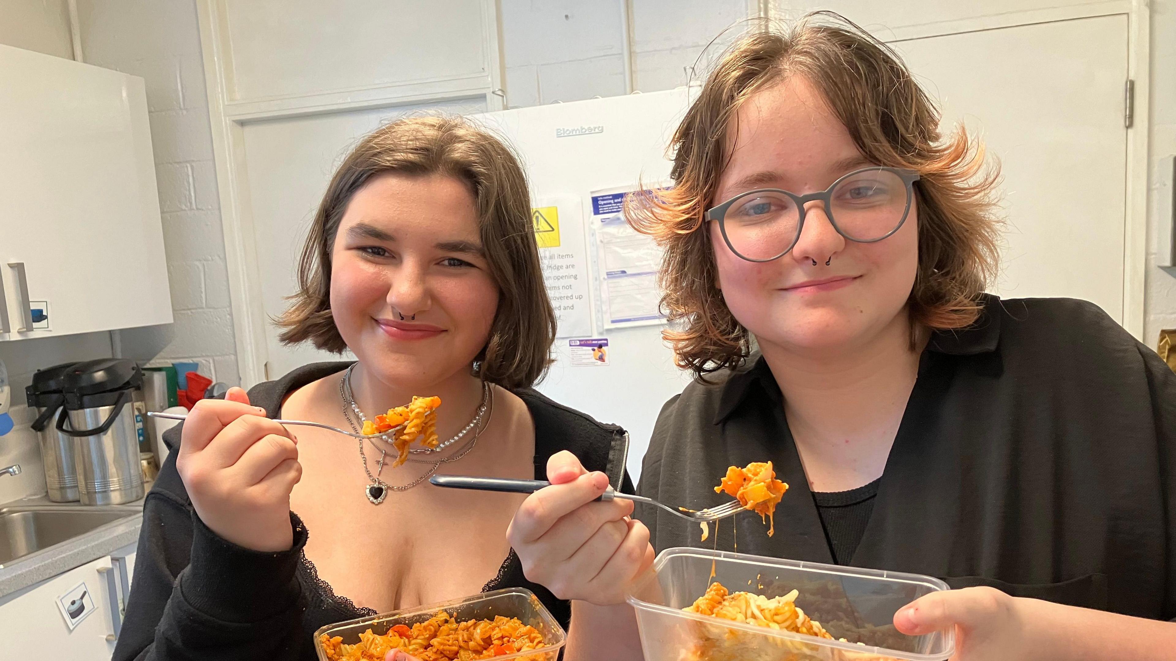 A picture of Lilli (left) and Scarlet (right) pictured holding two pots of take-aways. They both have brown hair and are smiling at the camera.