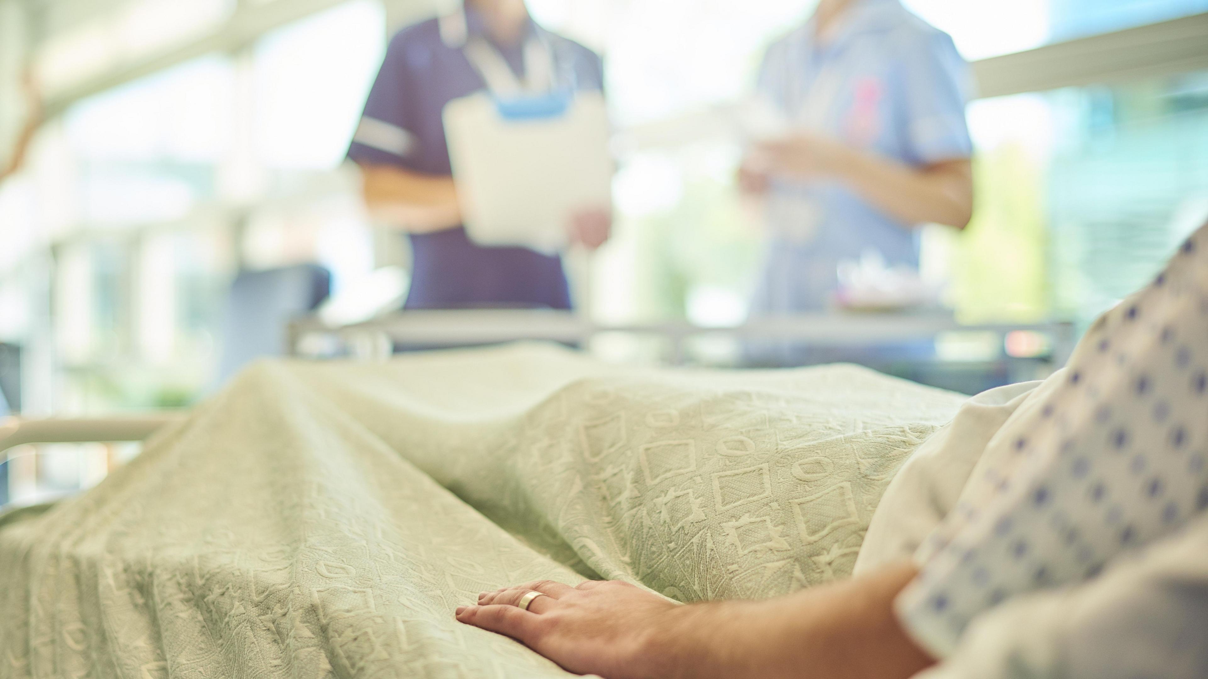 Man with a wedding band on his left hand wearing a hospital gown and lying in a hospital bed. Two nurses can be seen by the bed with a clipboard. 