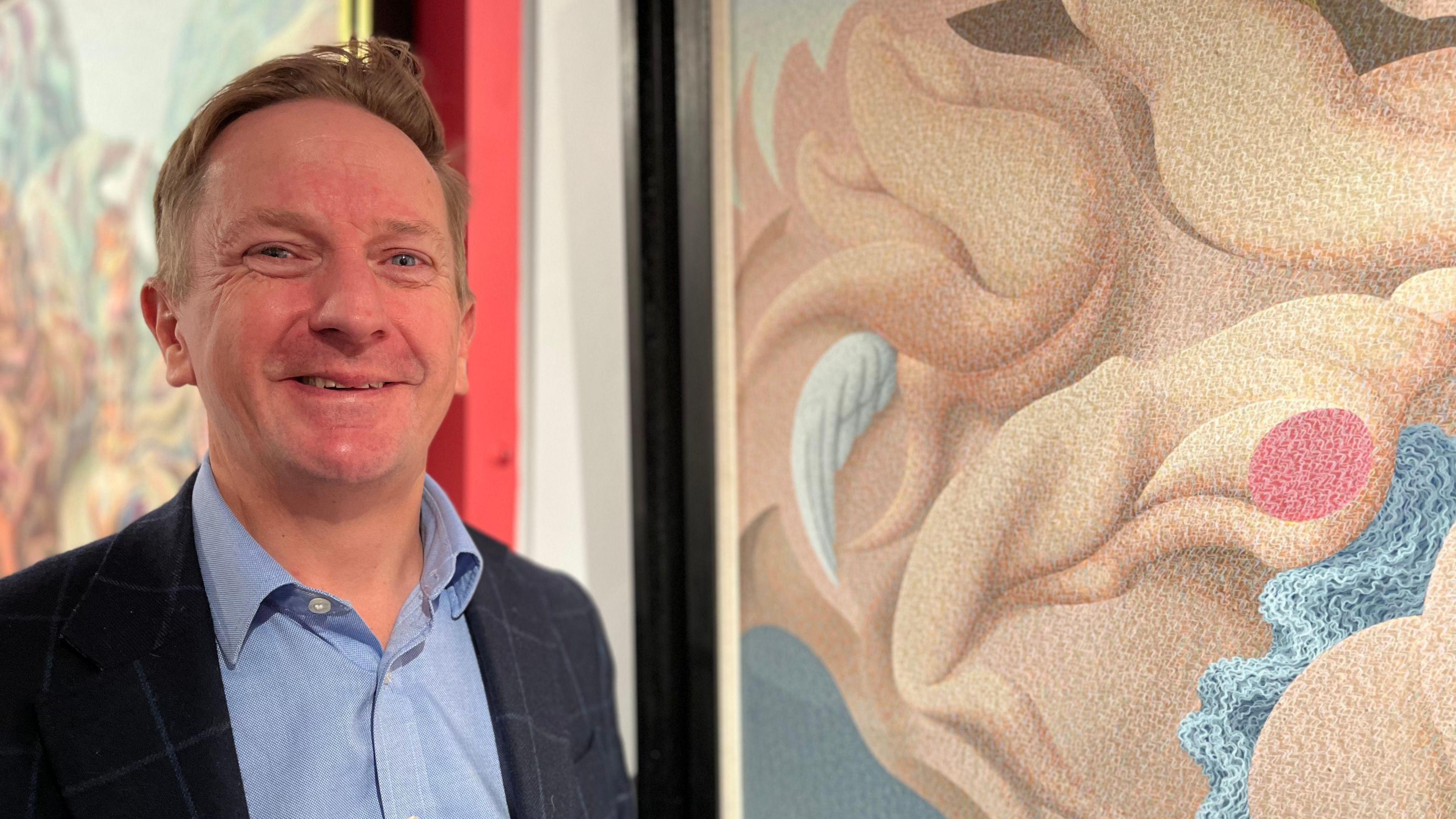 Grant Ford smiles at the camera wearing a blue shirt and check dark blue jacket. He stands in front of a surrealist canvas which has stylised forms of human forms melding into each other a wing in the mix. It's called Confusion.