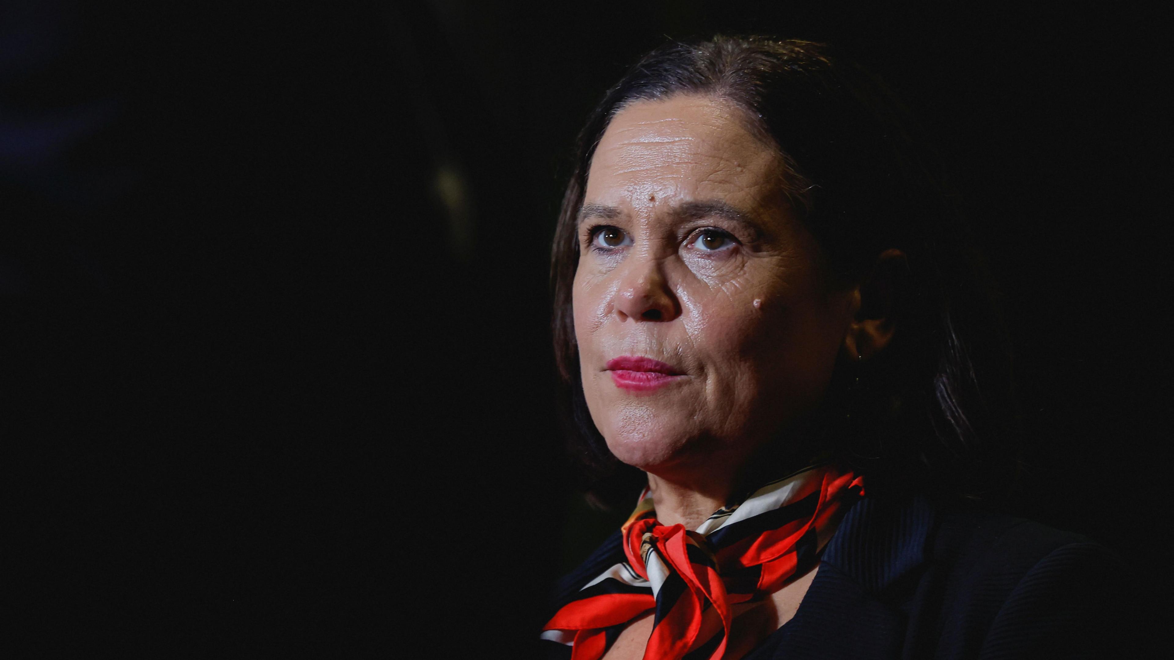 Mary Lou McDonald, pictured from the chest up, wearing a red scarf and jacket. The background of the image is dark.