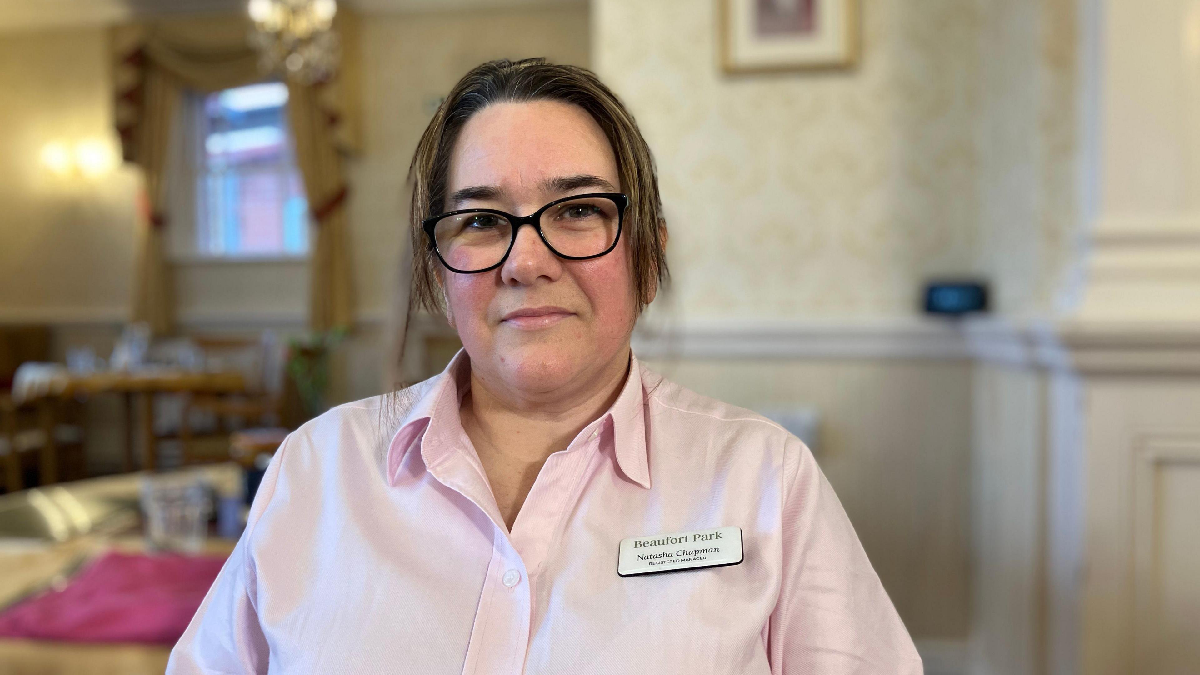 Natasha Chapman is sitting in the front room of a care home. She is wearing a light pink shirt and glasses.