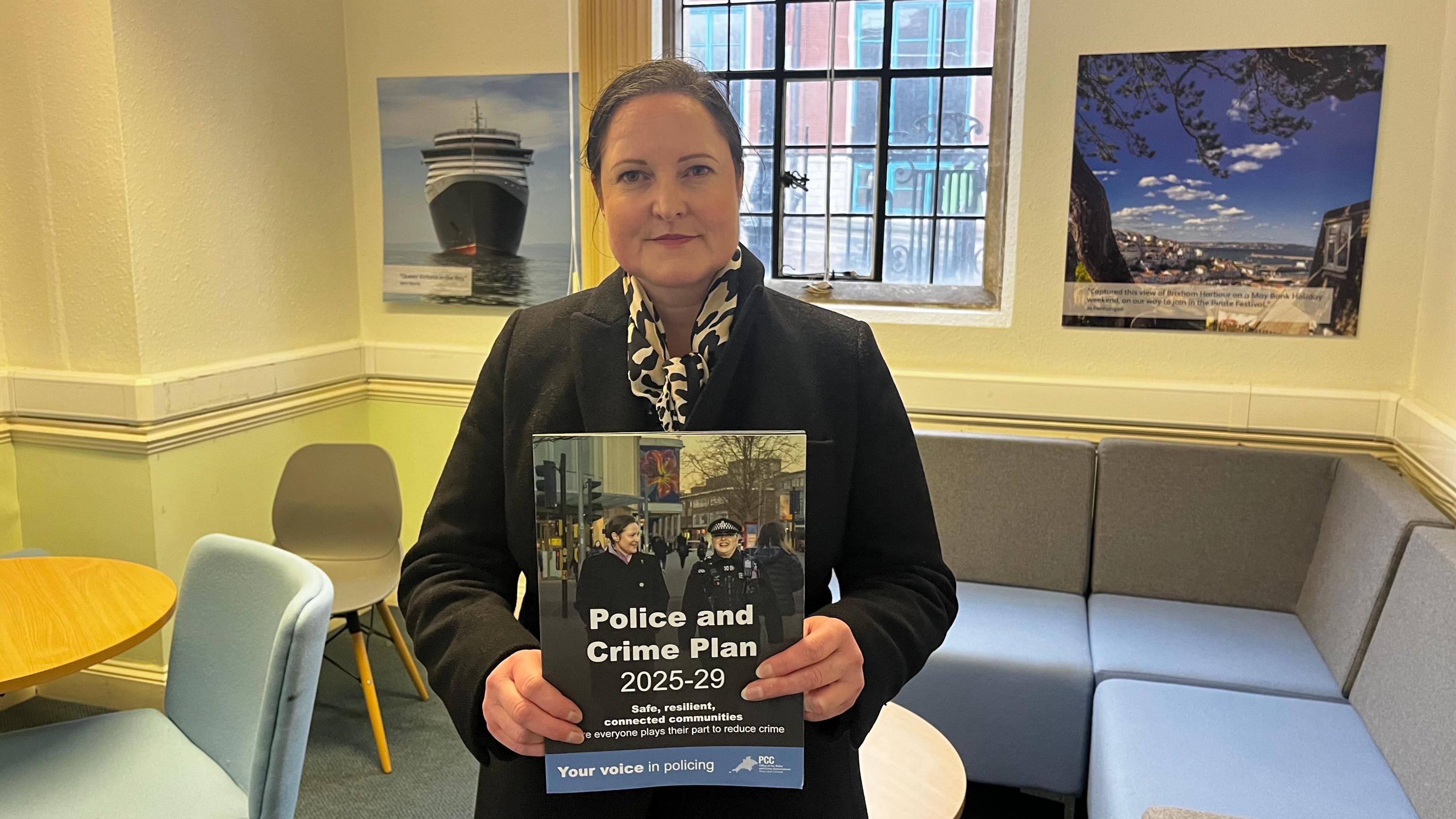 Alison Hernandez holds a booklet that says 'police and crime plan 2025-29'. She is inside an office room and is wearing a black coat and patterned scarf.