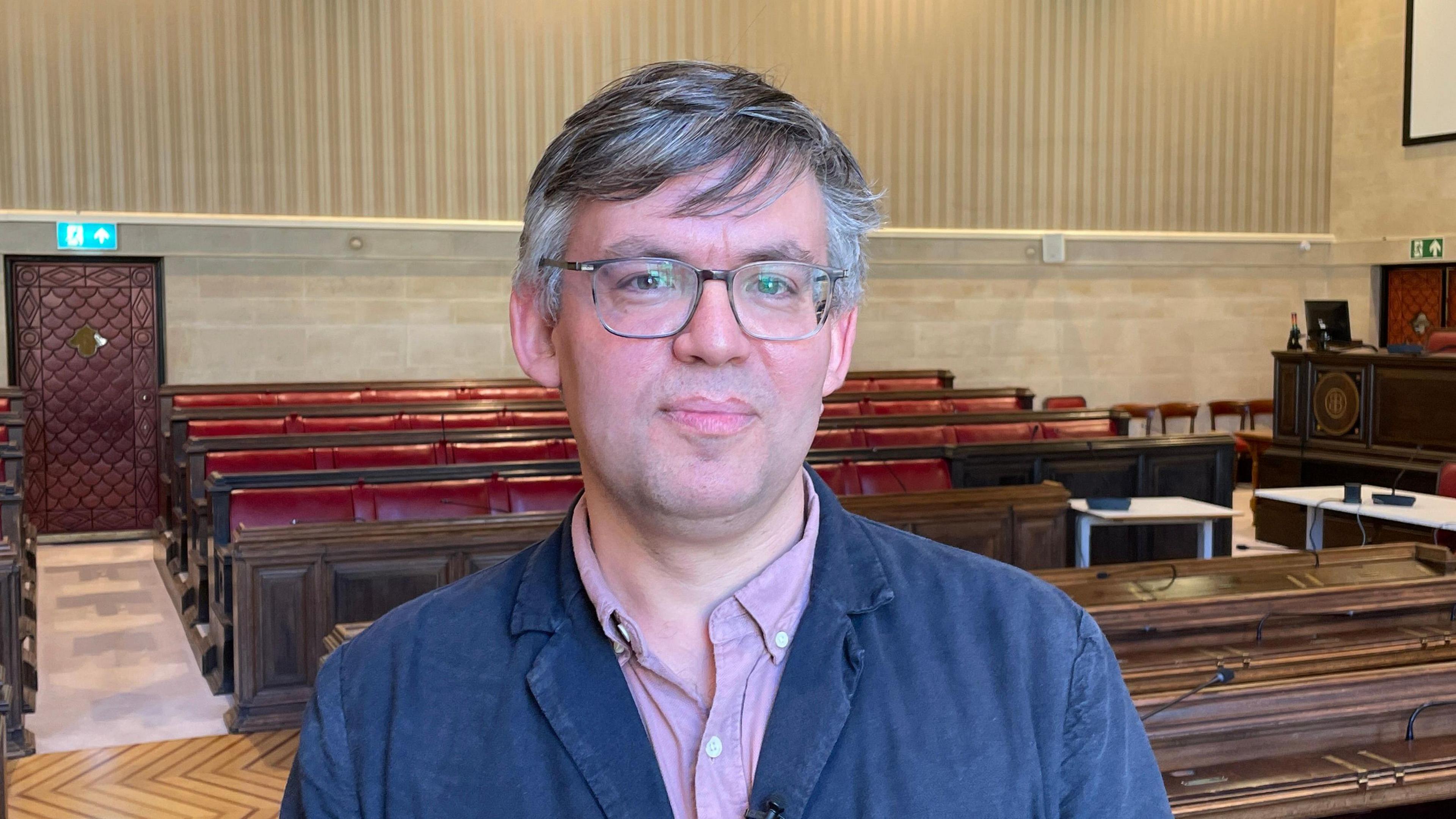 Barry Parson smiles at the camera. He is standing in a council chambers, with wooden pews seen behind him. He is wearing a dark blue jacket and a light purple shirt. He is wearing glasses. 