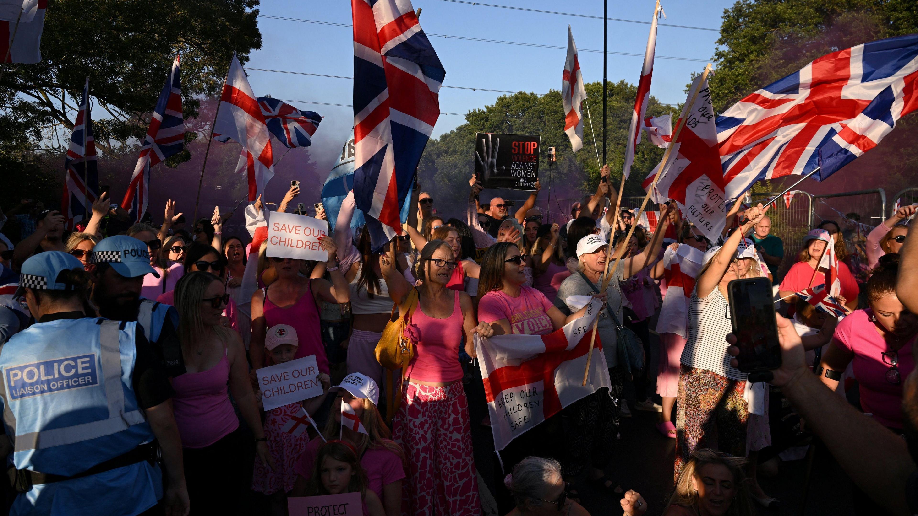 A large group of people, mostly women, take part in a protest. Lots of them are wearing pink and are holding the St George flag or Union Jack. Some are holding posters which say "save our children". 