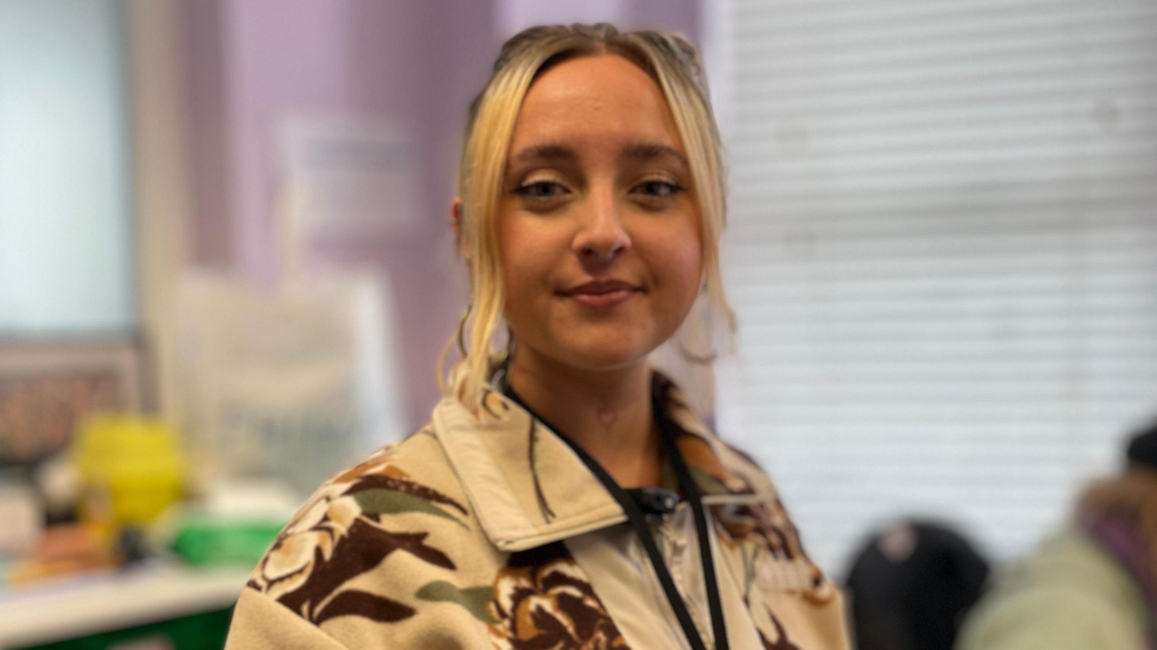 Keziah Cussen has blonde hair which is tied up. She is wearing a cream jacket with a foliage pattern on it. She is standing in front of a blind.