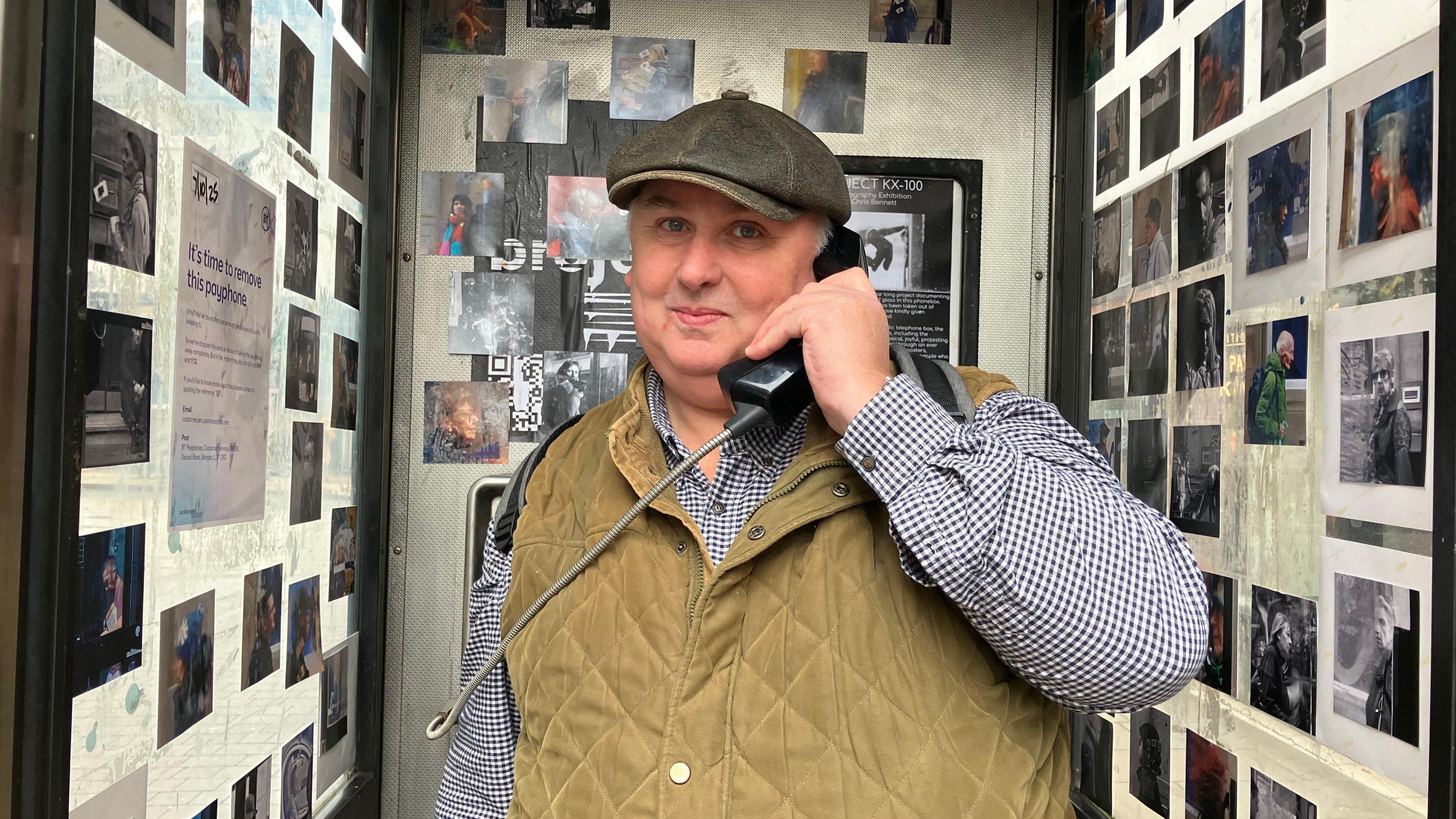 A man in a stone gilet, checked shirt and hat smiles as he holds a phone box phone to his ear. Behind him and to each sides are photos stuck onto the walls of the phone box.