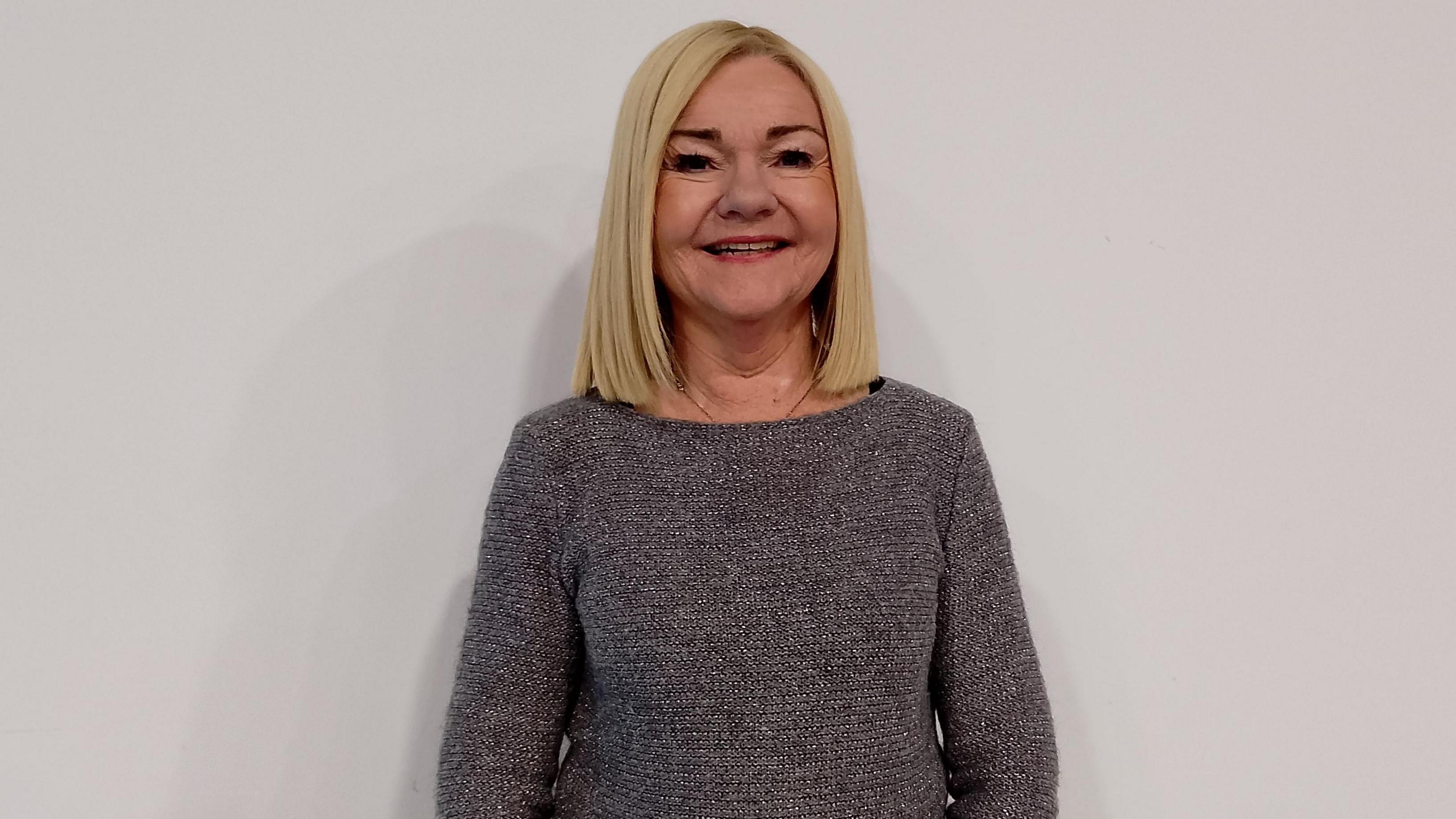 Paula Gibson is standing against a white wall. She's wearing a grey jumper and has a chain under her jumper.