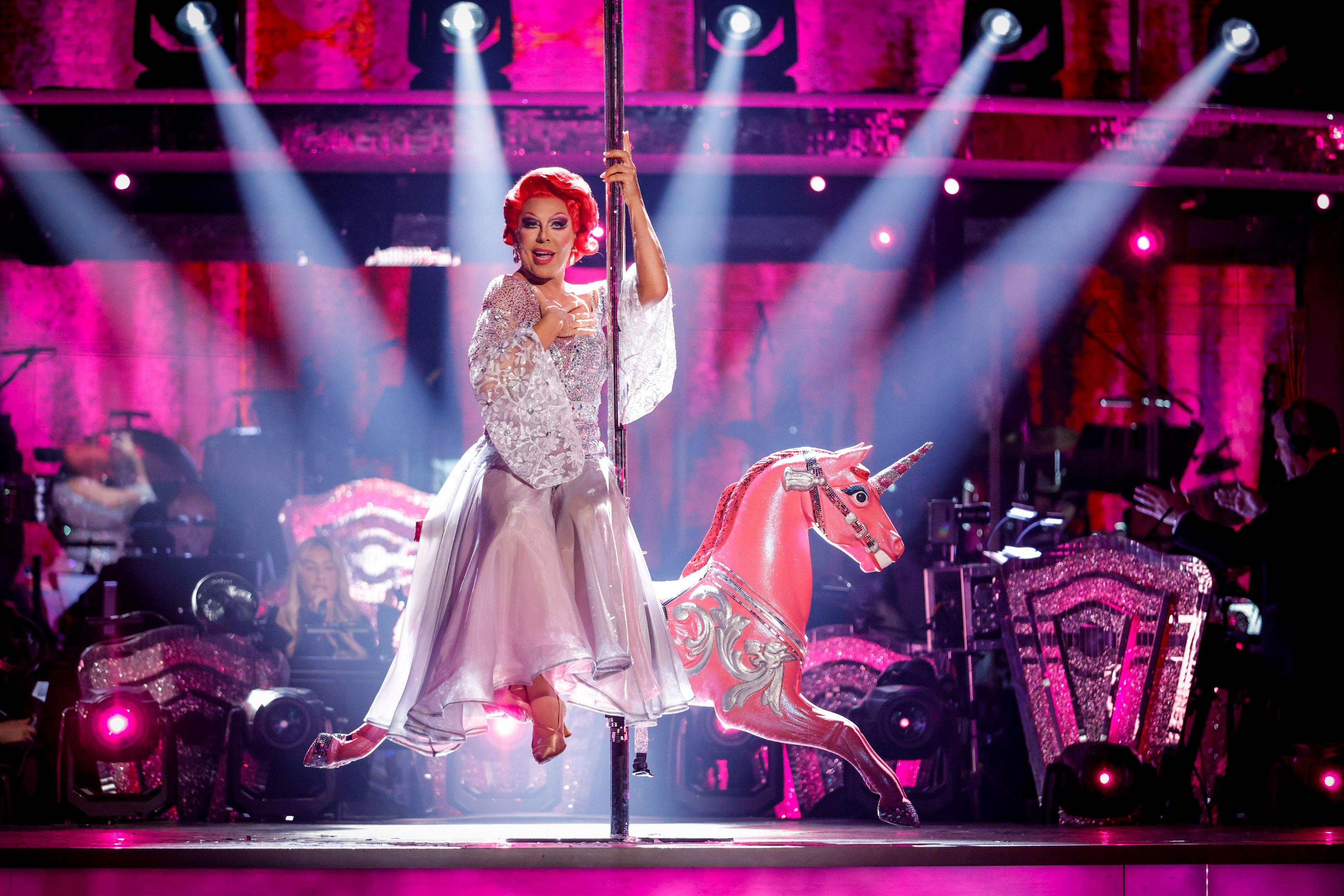 La Voix is on stage at Strictly Come Dancing, illuminated by a spotlight as she sits on the back of a carousel horse, holding its pole.