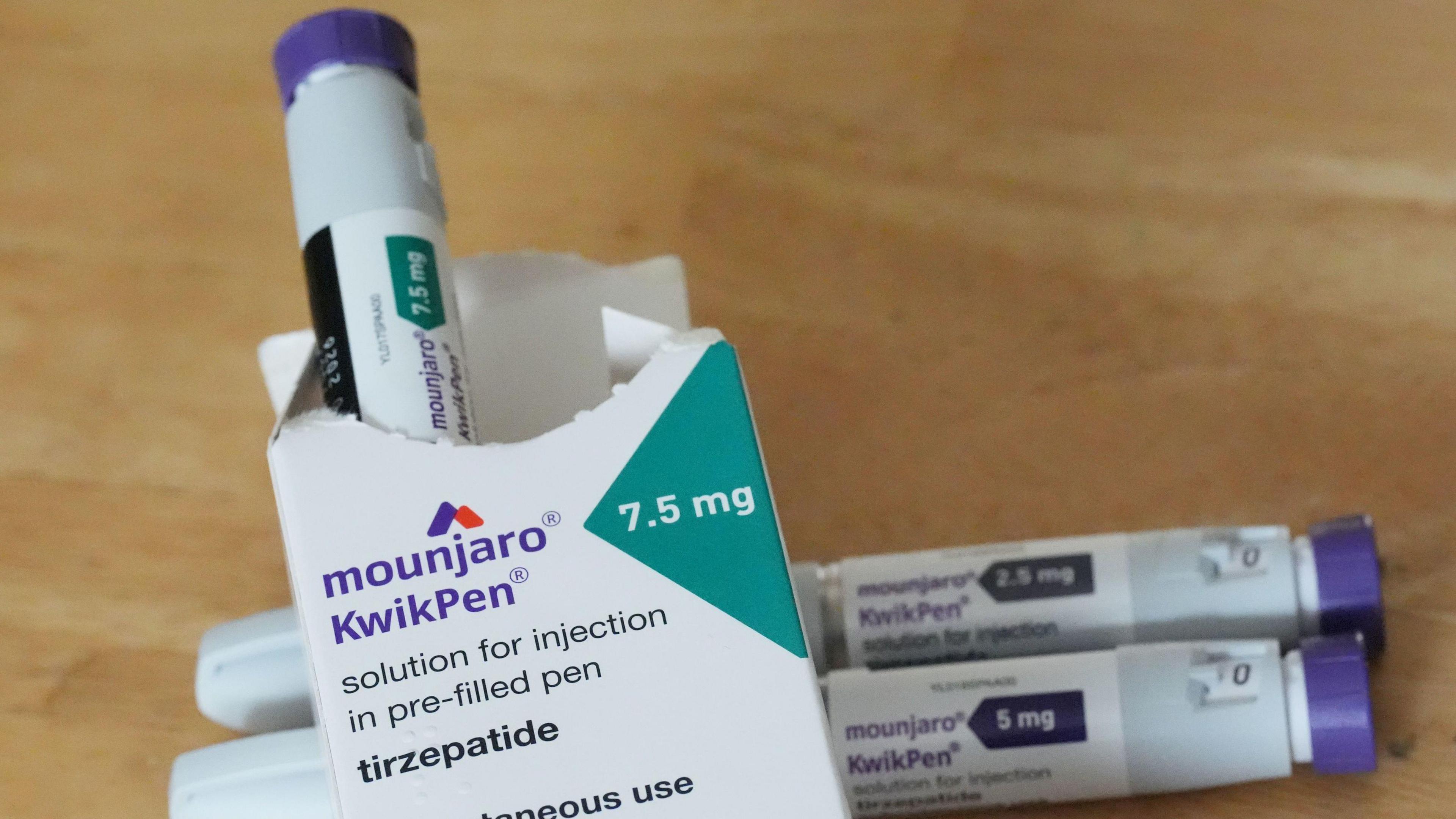 Pre-filled injection pens of 2.5mg, 5mg and 7.5mg doses of Mounjaro, the tirzepatide weight loss medication manufactured by US manufacturer Eli Lilly. One is coming out of the box and two are underneath on a wooden table.