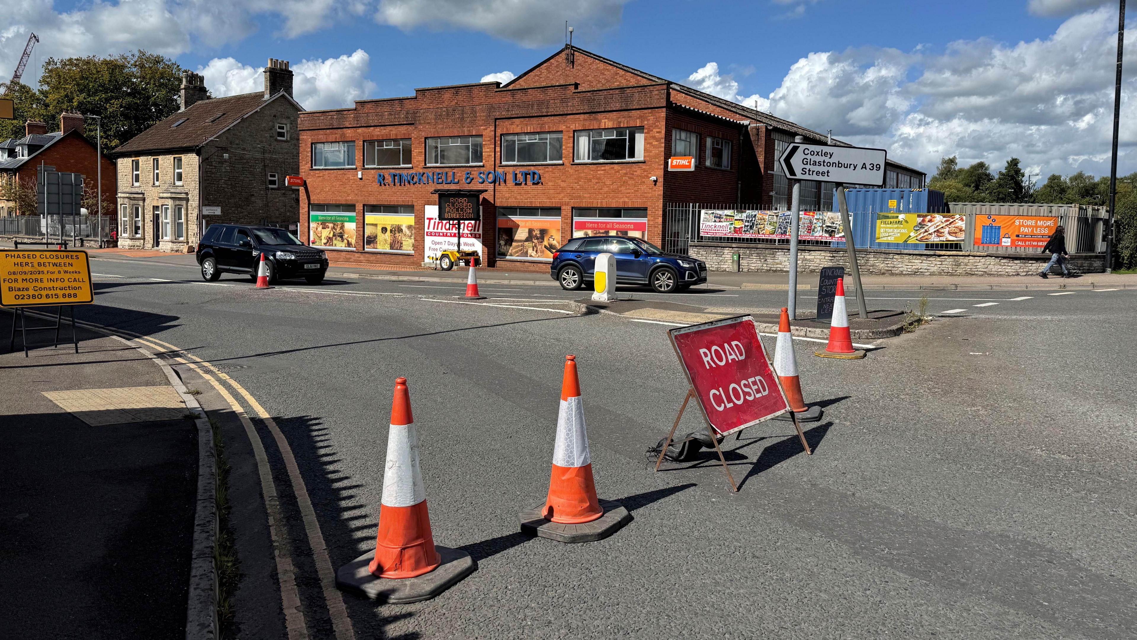 Four traffic cones and a big red ROAD CLOSED sign on the edge of a roundabout. There's a white sign pointing right saying Coxley, Glastonbury and Street on the A39. In the background there's a big bricked building and traffic is flowing onto the roundabout on the opposite side.
