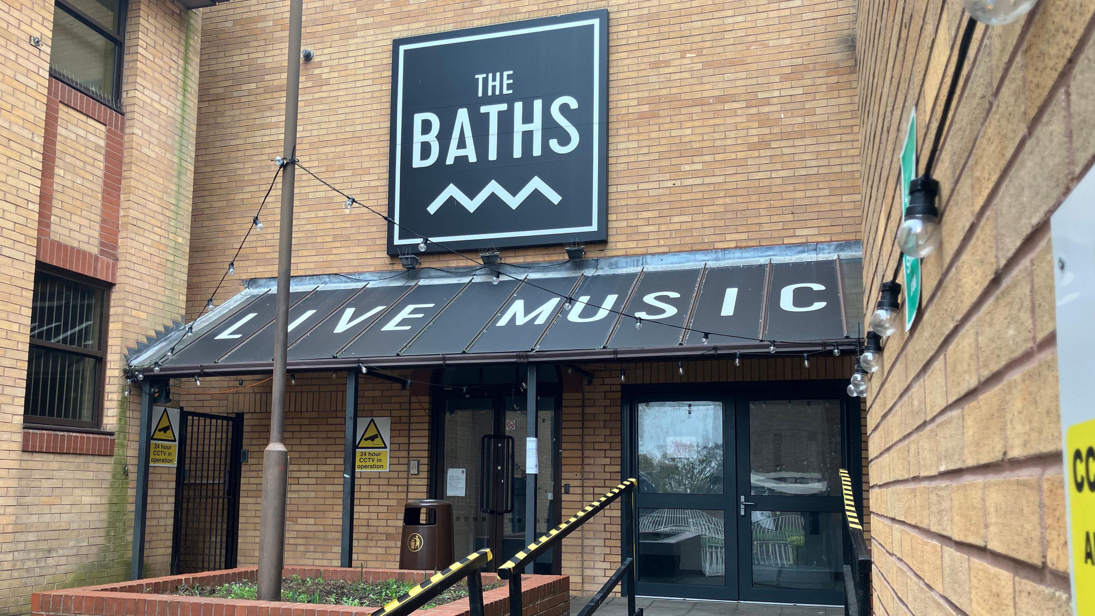 A general view of the entrance to The Baths in Ipswich. A black sign above the entrance reads 'The Baths' in white writing with a cover over the doors that reads 'Live Music' in white writing.