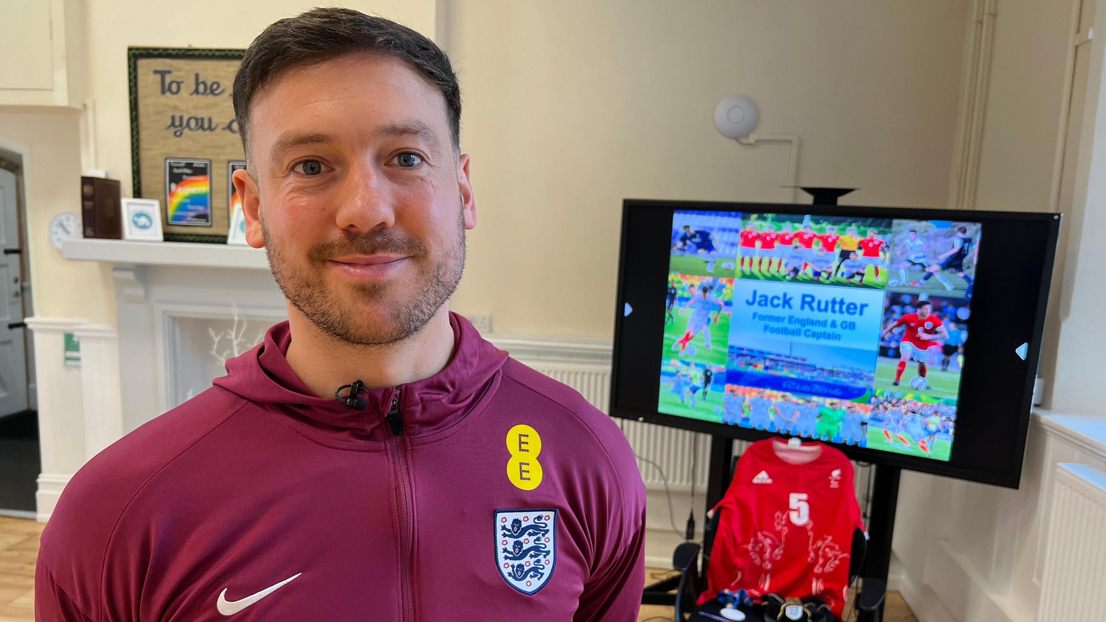 Jack Rutter, a man with brown hair that is very short on the sides, wearing a burgundy Nike hoodie, stands in a school hall. There is a projector screen behind him with a number of photographs from his sporting career, with text that reads "Jack Rutter, Former England and Great Britain Football Captain".