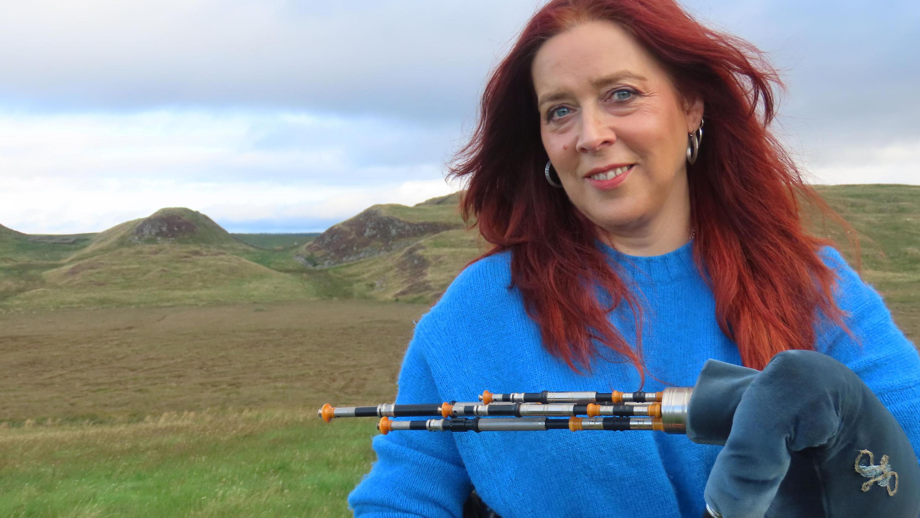 Head and shoulder shot of a smiling woman with red hair, wearing a blue jumper and holding a set of Northumbrian pipes. Behind her can be viewed moorland and rolling hills