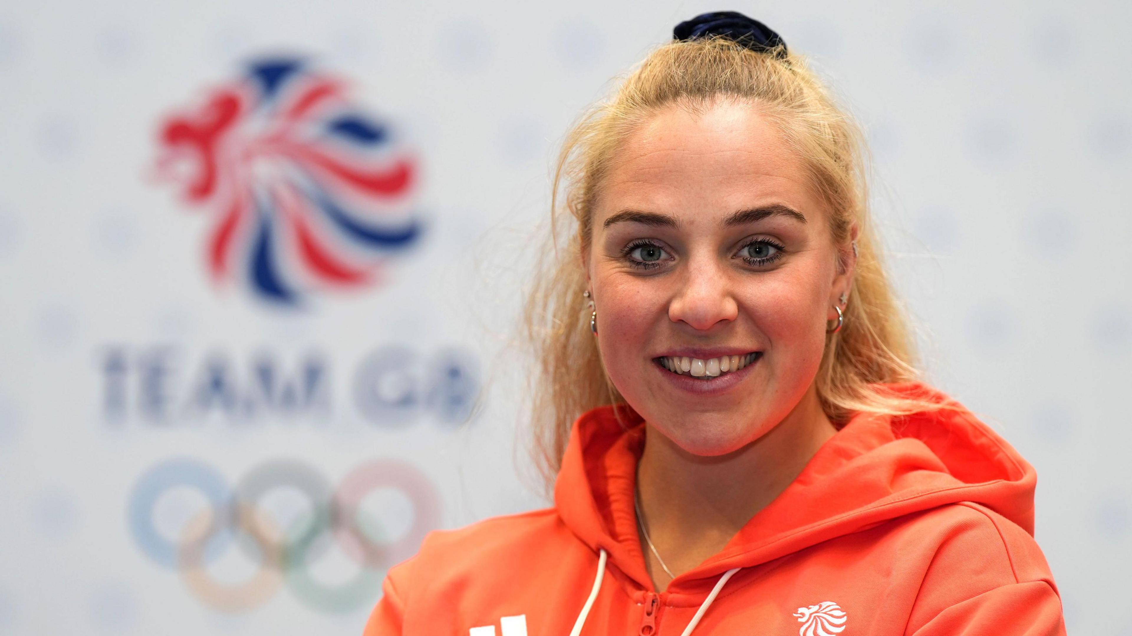 Becky Wilde smiling into the camera with the Team GB symbol blurred behind her and wearing an orange GB hoodie