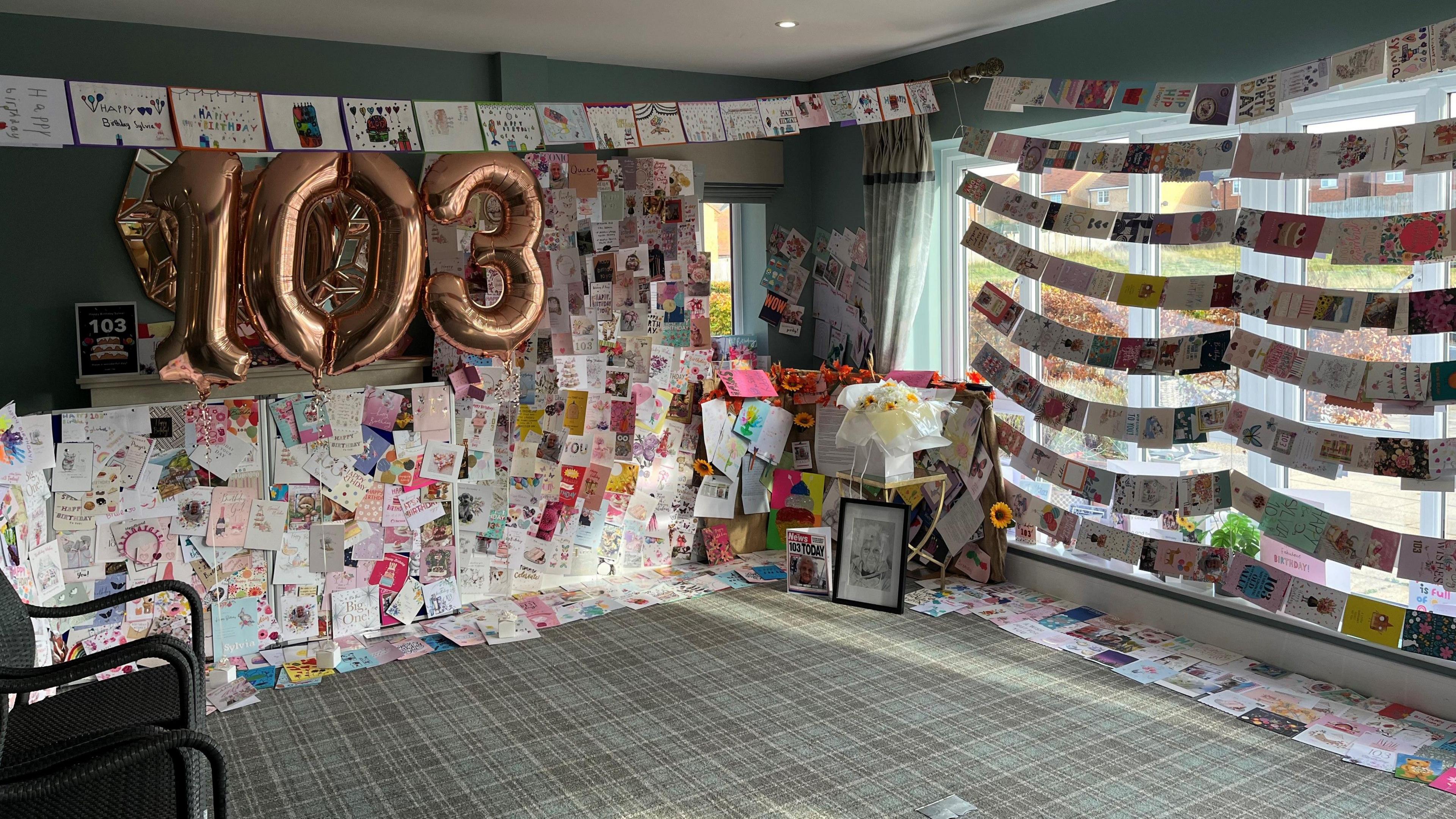 A carpeted room which is covered from floor to ceiling with birthday cards. On one wall the number 103 is hanging.