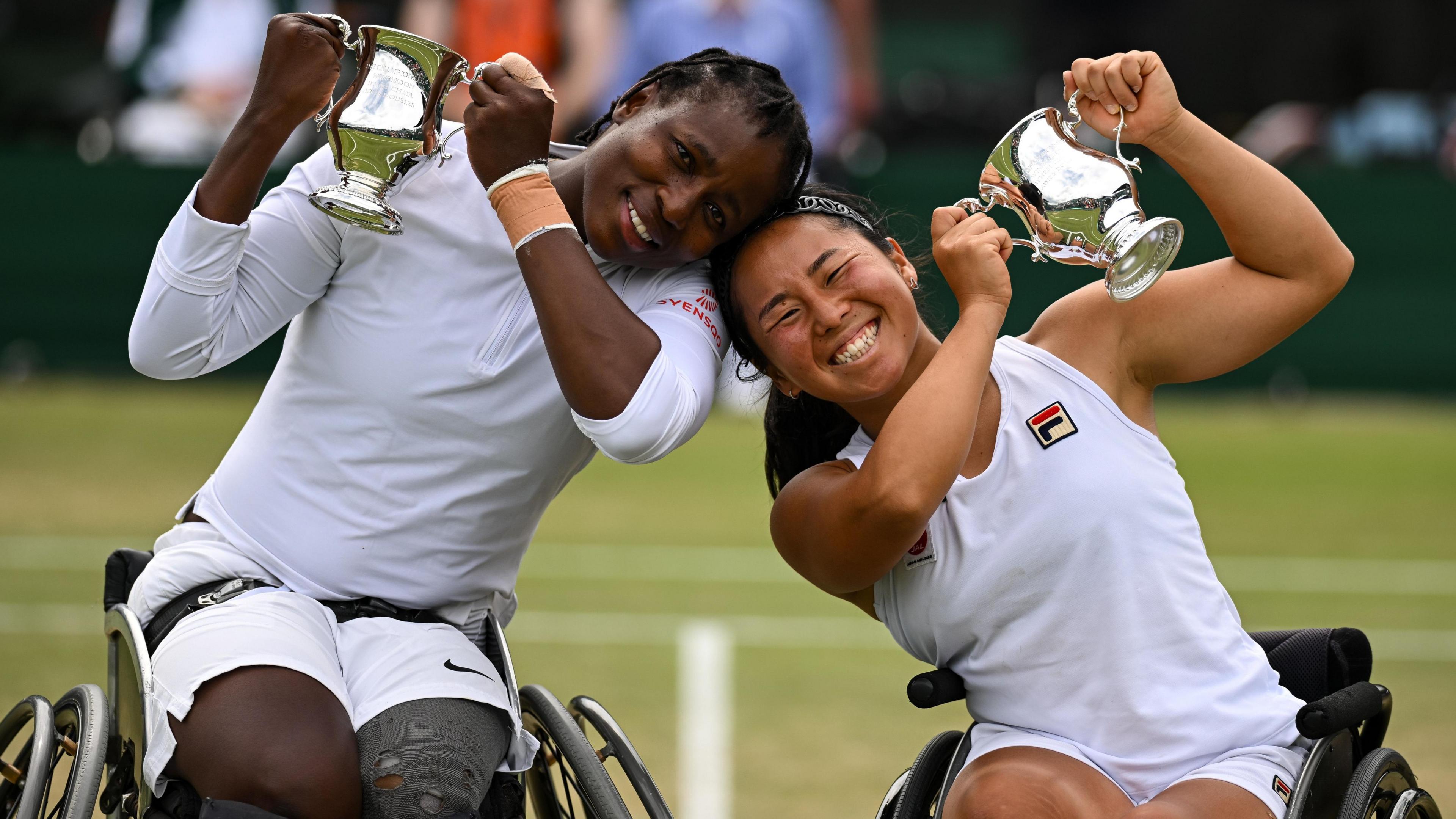 Kgothatso Montjane and Yui Kamiji, wearing tennis whites and sitting in wheelchairs, smile at the camera while holding aloft trophies after winning the wheelchair tennis women's doubles at Wimbledon