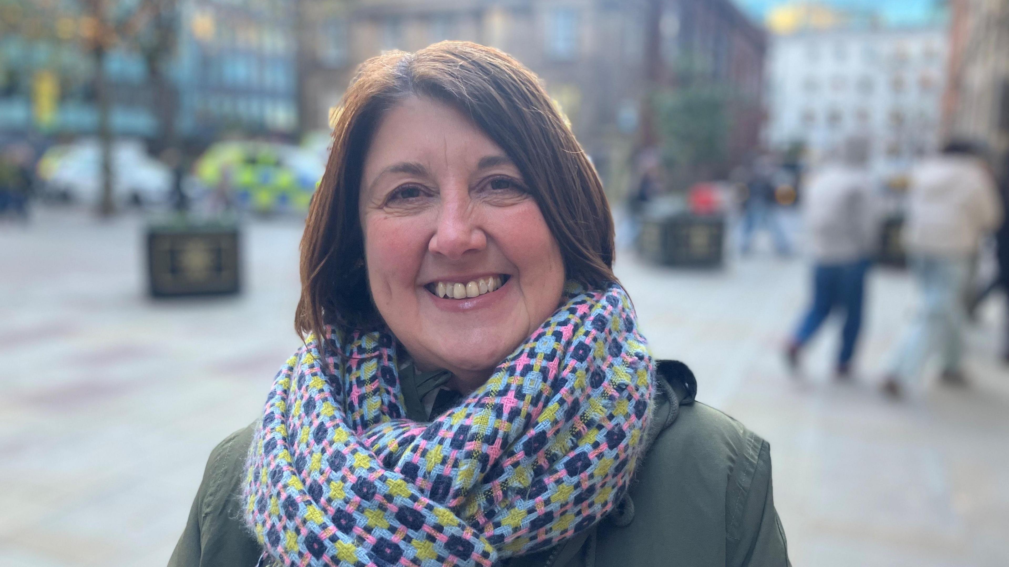 Photograph of Joanna Midgley, the Deputy Leader of Manchester City Council. She is pictured in Manchester city centre wearing a grey coat and large multi-coloured scarf.