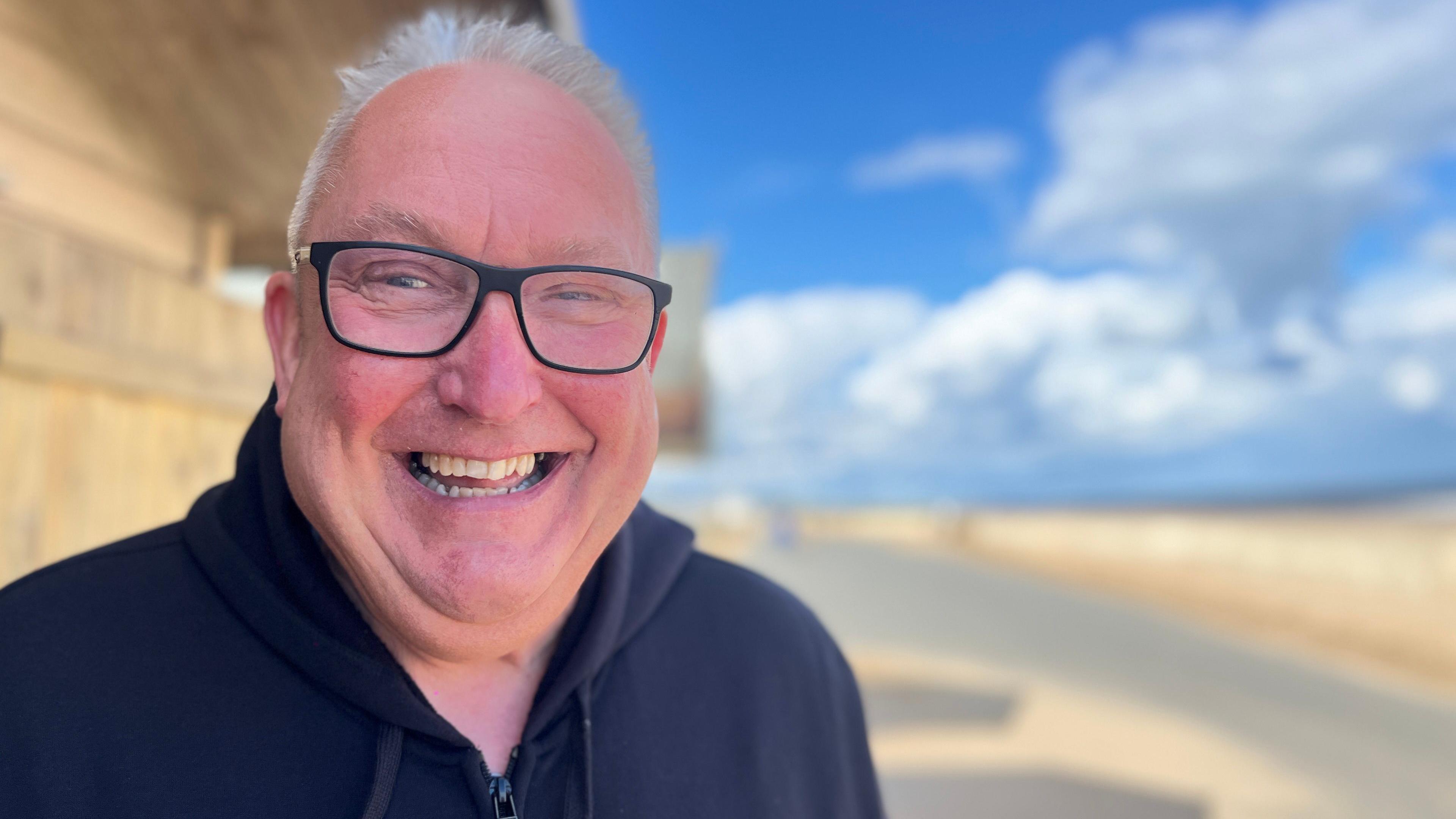 Clive Leeson smiles into the camera. He has short, grey hair and is wearing a navy hooded zip-up top and dark-rimmed glasses.