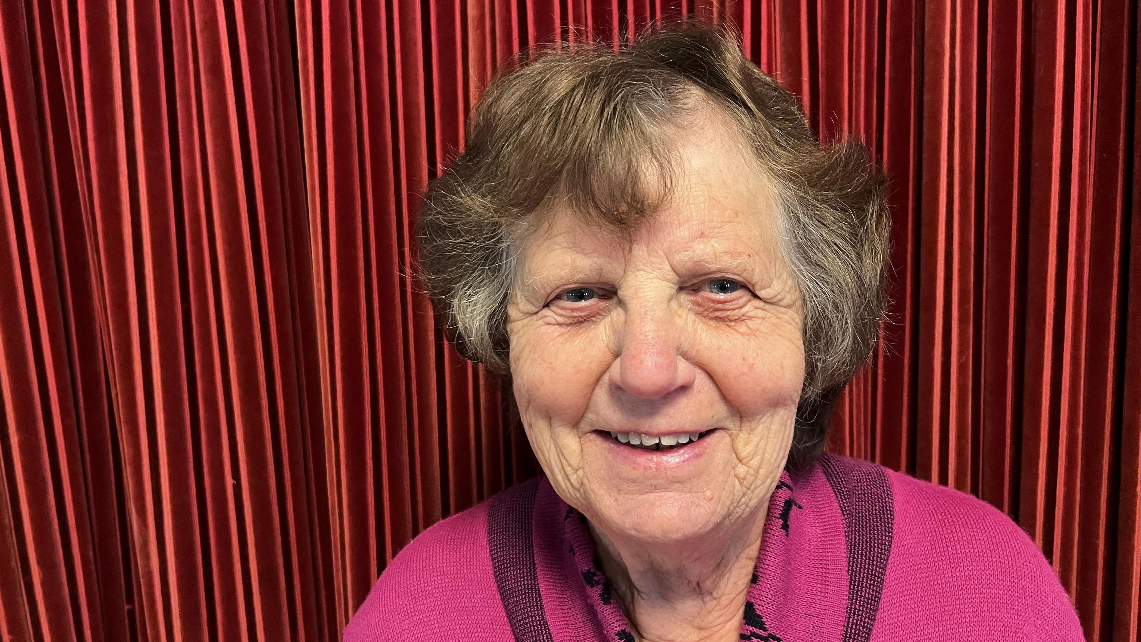 Janet Tweed, 83, has brown permed hair, with some grey flecks, she is smiling and is wearing a pink woollen top with black patterns in it. behind her are some red velvet curtains of a stage.
